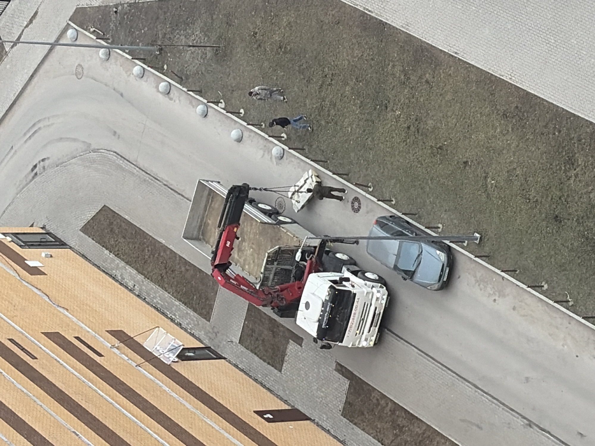 The image depicts an outdoor scene in a modern residential area of Saint Petersburg, Russia, observed from a high vantage point under overcast daylight conditions.
A white heavy-duty truck, identified as a "МАНИПУЛЯТОР" (truck-mounted crane) with a red hydraulic crane arm, is positioned on a narrow, asphalt road. The truck's cab displays the partial phone number "+7 912 1448-00-44". Its crane arm is actively lowering a large, rectangular, light-colored object, possibly a packaged building component, wrapped in protective material.
One male individual, dressed in dark clothing, stands directly beneath the suspended object, extending his arms to guide its descent onto the paved area. Two additional male individuals are present on the adjacent grassy strip to the right: one in a dark jacket and blue jeans, walking towards the activity, and another further back in a lighter jacket and pants, also observing.
A dark grey/blue Ford Focus sedan is parked on the asphalt road directly in front of the truck, facing away from the camera. Its rear license plate is affixed but illegible.
To the left of the road is a multi-story building featuring a facade of light brown/orange bricks with prominent horizontal dark brown bands. Several dark-colored entrance doors are visible, one covered by a clear, sloped canopy. A light grey paver stone sidewalk runs between the building and the road.
To the right of the road, a strip of sparse, dull green/brown grass is delineated by a line of light grey spherical concrete bollards and thin, dark posts. The asphalt road itself shows tire marks and contains at least two circular manhole covers. A dark-colored lamppost stands on the right side of the road, near the grassy verge.