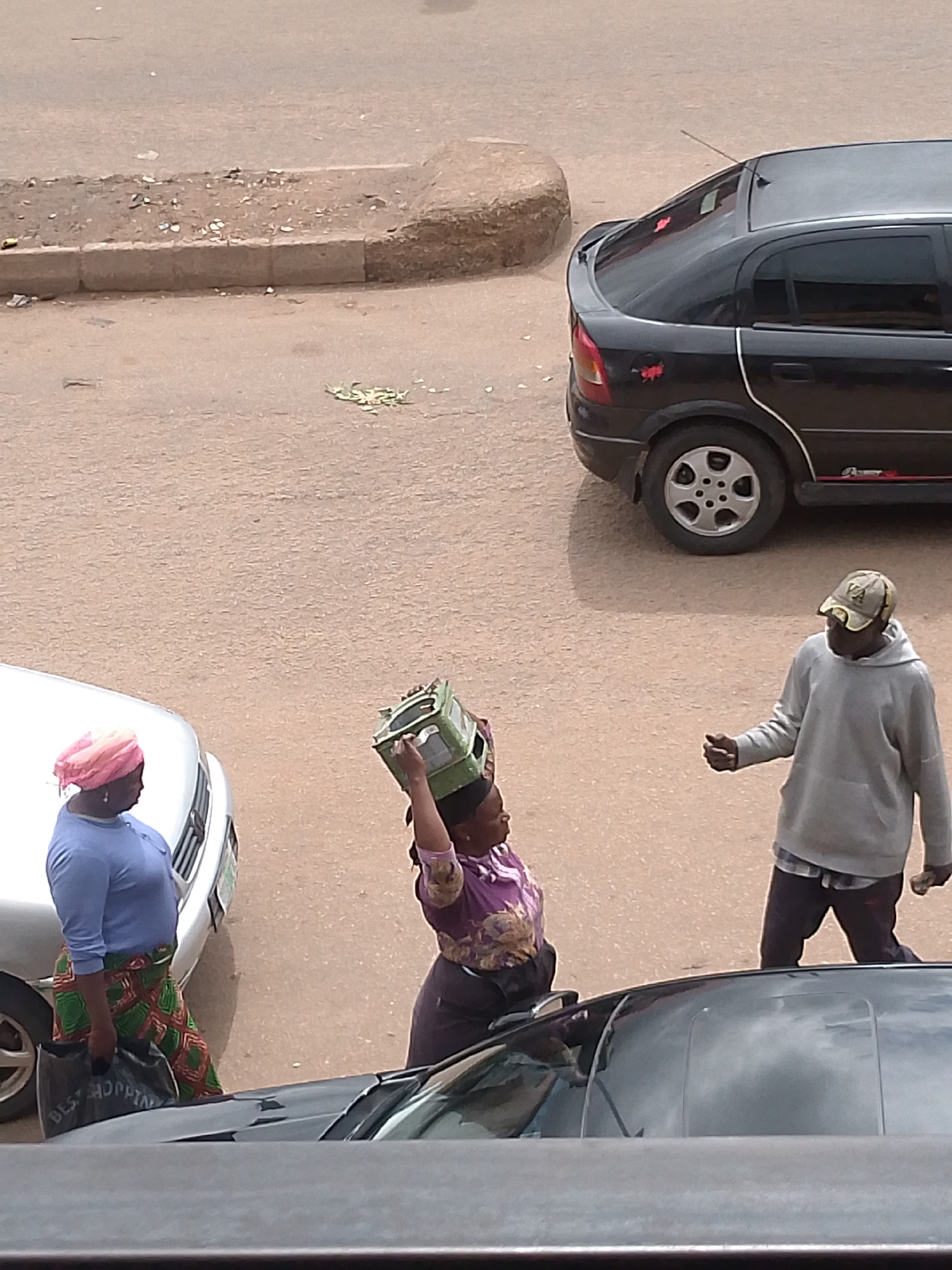 In Bukuru, Nigeria, a woman wearing a pink headscarf and a blue shirt, along with a green and red patterned skirt, stands next to a white car. She is carrying a black shopping bag. To her right, another woman, dressed in a purple and gold patterned top and a dark skirt, balances a green crate on her head. A man wearing a light gray hooded sweatshirt and a camouflage cap walks away from the woman with the crate, gesturing with his right hand. A black car is parked behind the man. The scene takes place on a paved street with a curb on the left side, where debris is scattered.