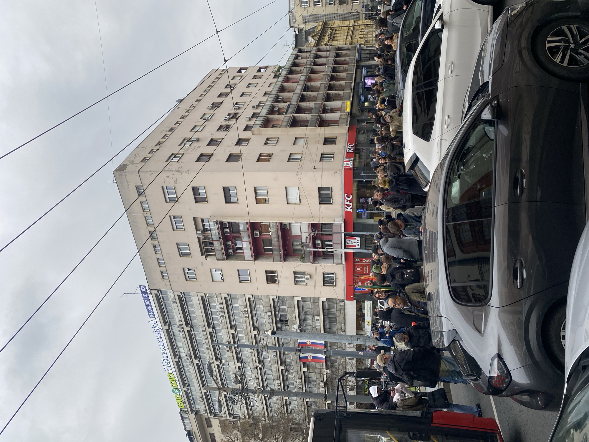 The image depicts a street scene in Belgrade, Serbia, characterized by a crowd of people gathered alongside a tall, aged building. The building features numerous windows and a KFC restaurant on the ground floor. Serbian flags are displayed on lampposts. A line of cars is partially visible on the right side of the image, and a bus is at the bottom, suggesting traffic congestion. The sky is overcast. People are queuing, likely for the KFC. The architectural style of the building suggests a post-war era.