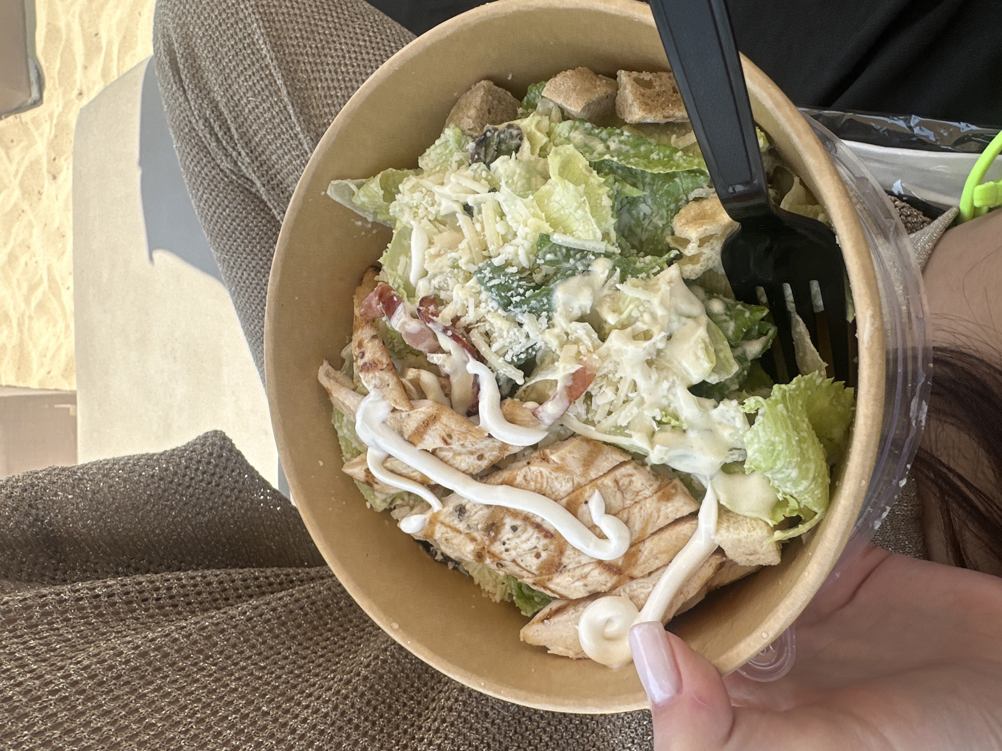 A person's hand with a light pink manicure is holding a disposable bowl of Caesar salad. The salad contains chopped grilled chicken, romaine lettuce, croutons, and shredded cheese, all coated in a creamy dressing. A black plastic fork is inserted into the salad. The bowl is made of light brown paper. In the background, a person is seated on a sun lounger, covered by a knitted brown blanket. The environment appears to be a beach or poolside area, with sand visible in the upper portion of the image and a portion of a resort umbrella casting a shadow.