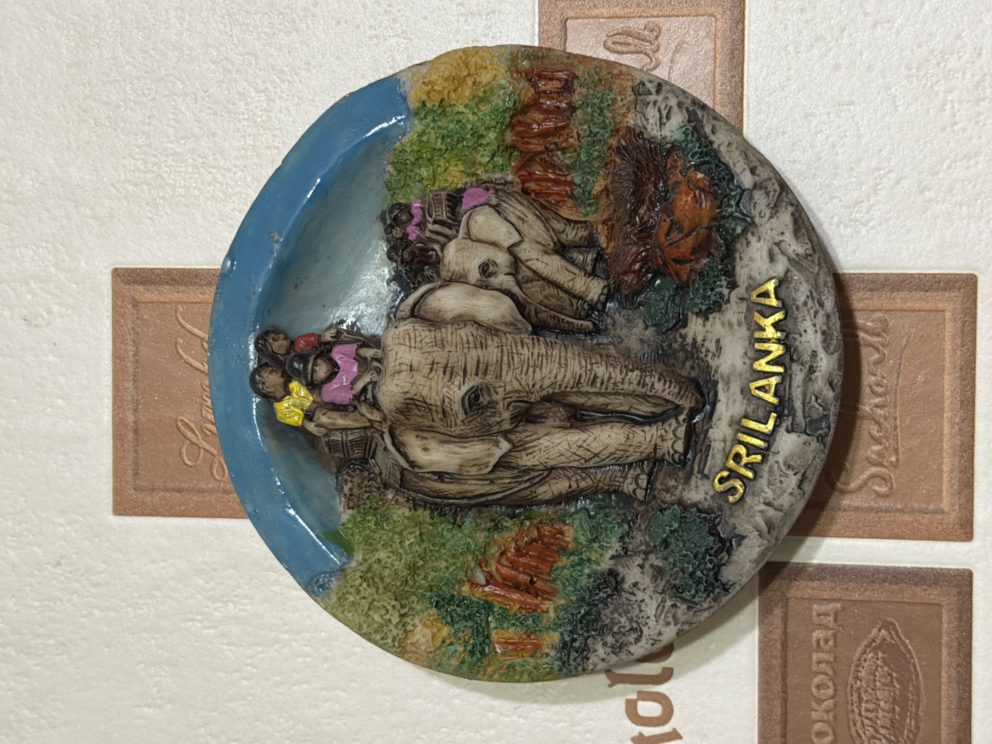 The image shows a souvenir plate or magnet depicting elephants and inscribed with "SRI LANKA" in gold letters. The plate has a blue rim, and the design includes details of a landscape with trees, rocks, and what appear to be two elephants. The plate is positioned against a wall with textured white wallpaper. Partially visible below the plate are two small brown tile-like objects with embossed text in a foreign language and a cocoa bean symbol. The scene suggests a location in Krasnoyarsk, Russia, based on the tile objects.
