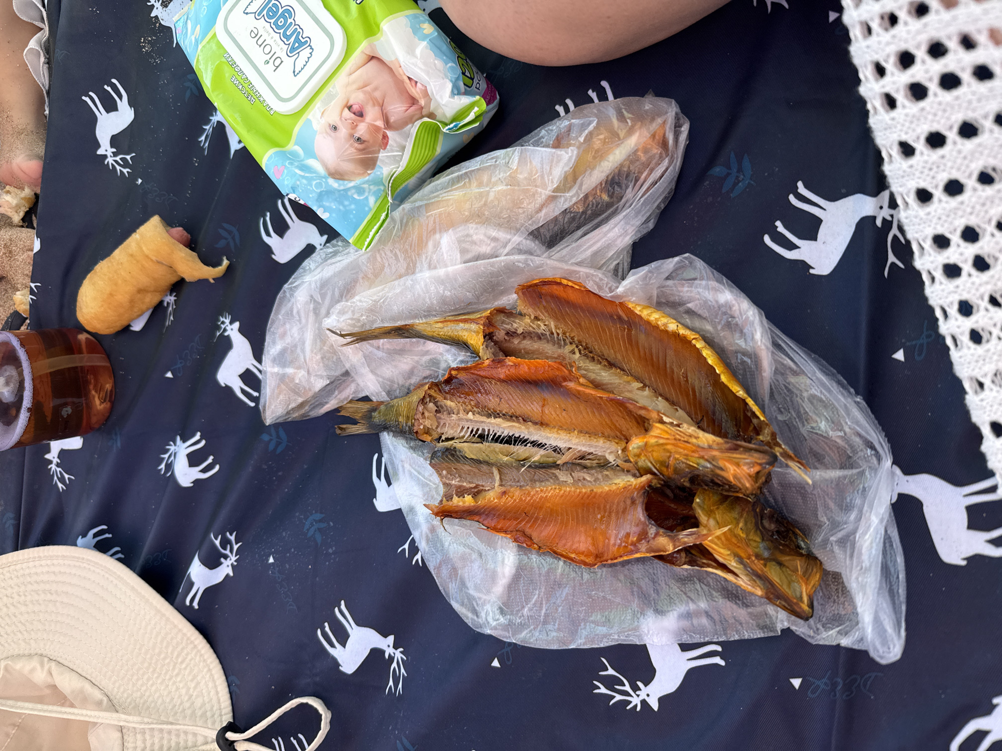 A close-up overhead shot shows food and items laid out on a dark blue picnic blanket with a white deer pattern. In the foreground, three smoked fish, split and cleaned, are arranged in a clear plastic bag. To the right of the fish, a packet of "bione Angel" baby wipes is visible. Further up and to the right, a piece of fried food, possibly a corn dog, lies on the blanket next to a glass of amber liquid. A light-colored hat with a drawstring is in the upper left corner, and a white crocheted edge is in the lower right. The setting appears to be a beach or outdoor picnic area, with sand visible in the upper portion of the image. The overall scene suggests a casual meal or snack. The location is identified as Sary-Oy, Kyrgyzstan.