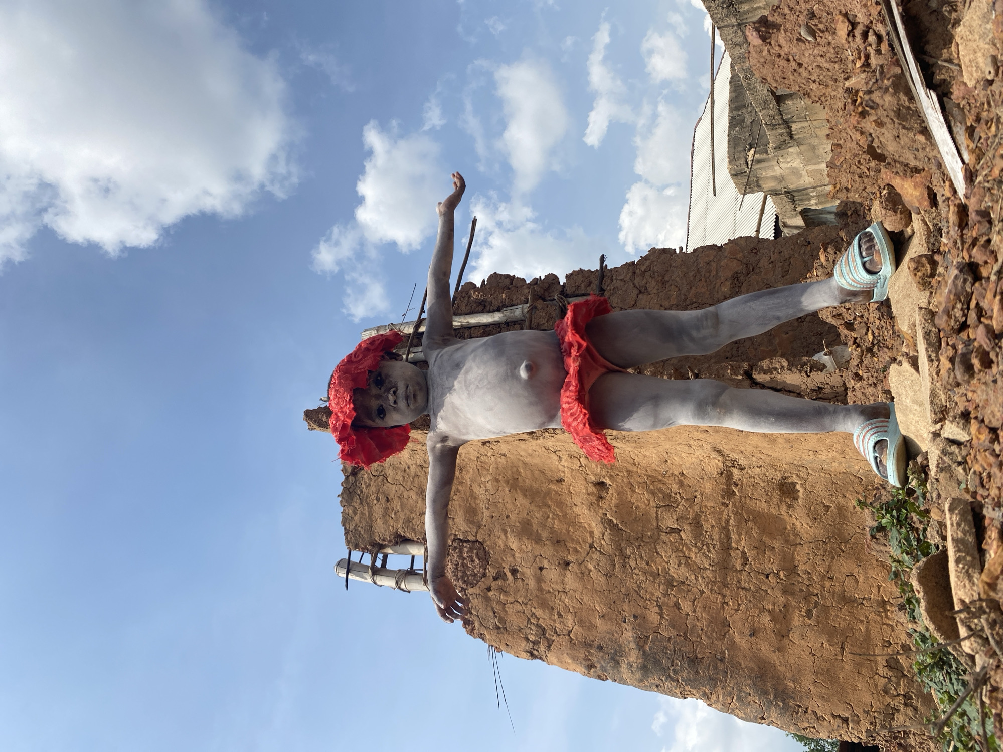A person covered in grey paint and wearing a red headpiece and red clothing is standing on top of a mud building in Igbarra, Nigeria. The person is wearing light blue sandals. The sky is visible in the background, and there are clouds.