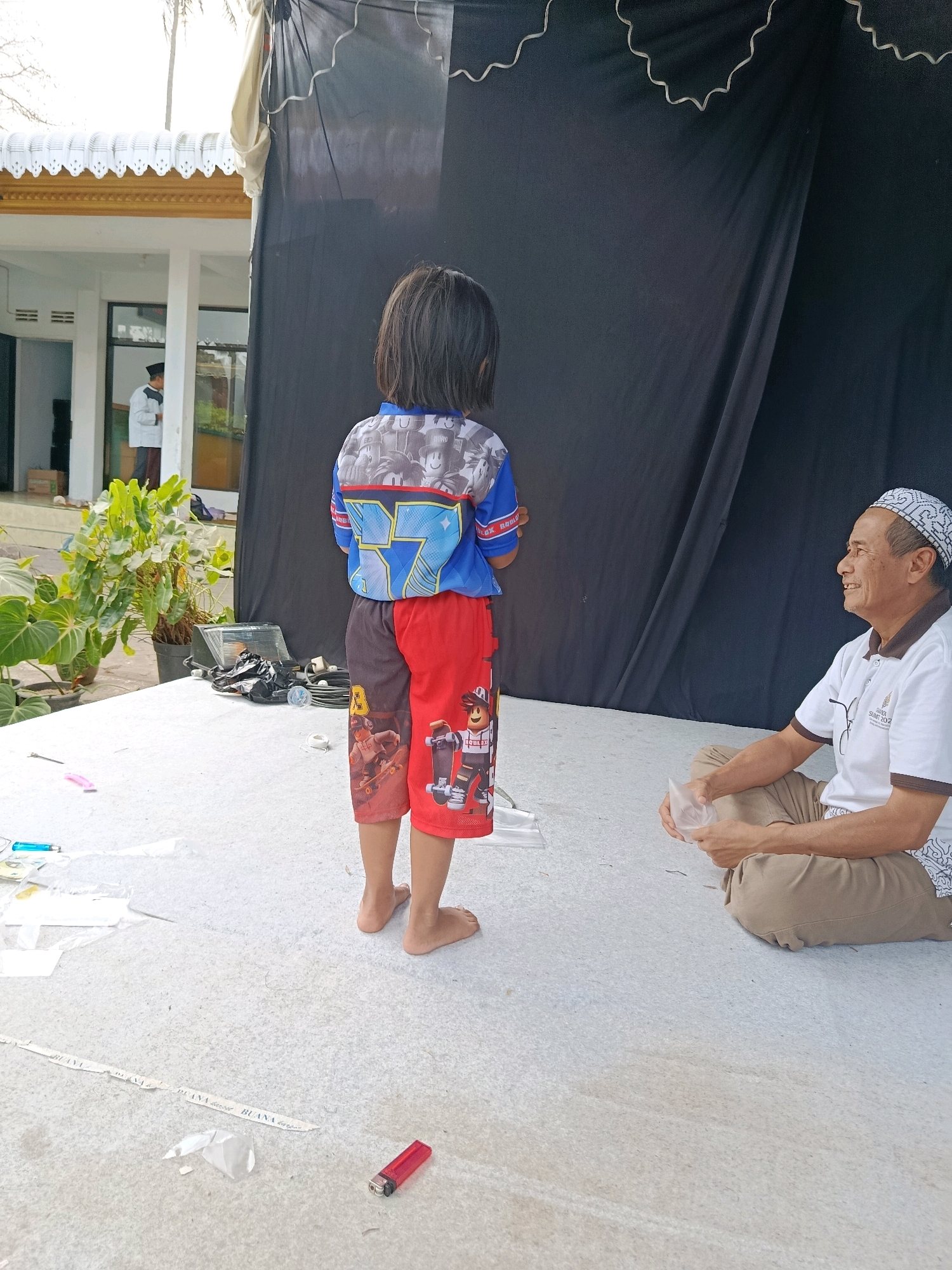 In Bululawang, Indonesia, a young child stands with their back to the camera, facing a black curtain. The child is wearing a blue and red shirt with cartoon characters and red shorts adorned with Roblox characters. To the right, an elderly man with a white and black patterned cap and a white polo shirt sits on the floor, holding a small plastic bag. He is looking towards the child and smiling. The ground is a light-colored, textured surface, possibly concrete or a similar material. In the background, a building with white pillars and decorative trim is visible, along with lush green plants. Various items, including what appears to be a lighter, plastic wrappers, and tape, are scattered on the ground. The scene suggests an outdoor or semi-outdoor setting, possibly a temporary stage or event area, with a backdrop set up for an activity or performance.