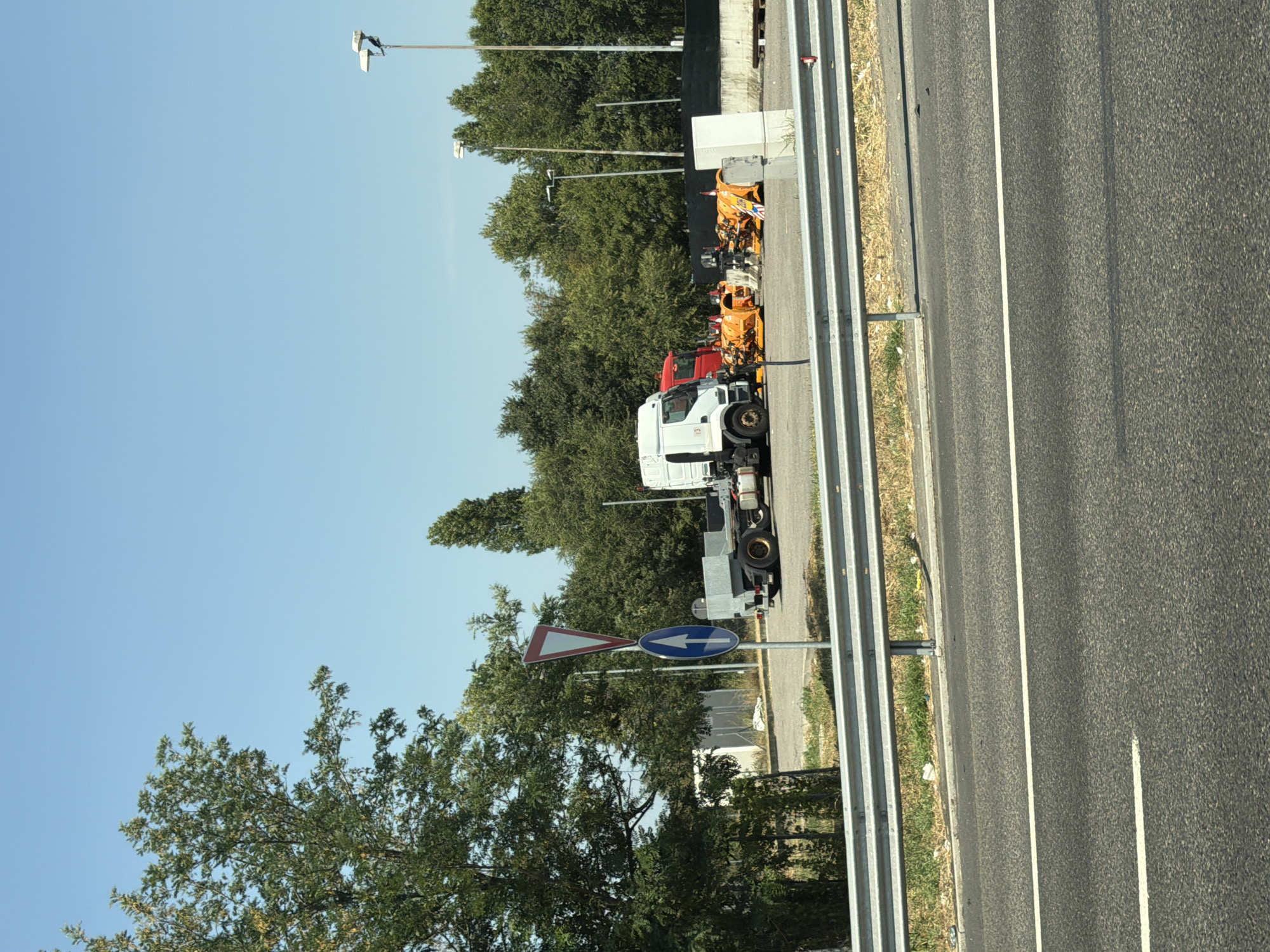 The image shows a road with a guardrail in the foreground, and a clear blue sky above. Behind the guardrail, there is a dirt lot with several trucks and equipment. A white truck with a trailer is parked in the lot, along with a red truck. Several orange snowplows are also visible, as well as a traffic sign with a red triangle and a blue circle with a white arrow. Trees and bushes line the background of the lot. The scene appears to be an industrial area or a storage yard. The location is identified as Lainate, Italy, suggesting this is likely a road or industrial area within that municipality.