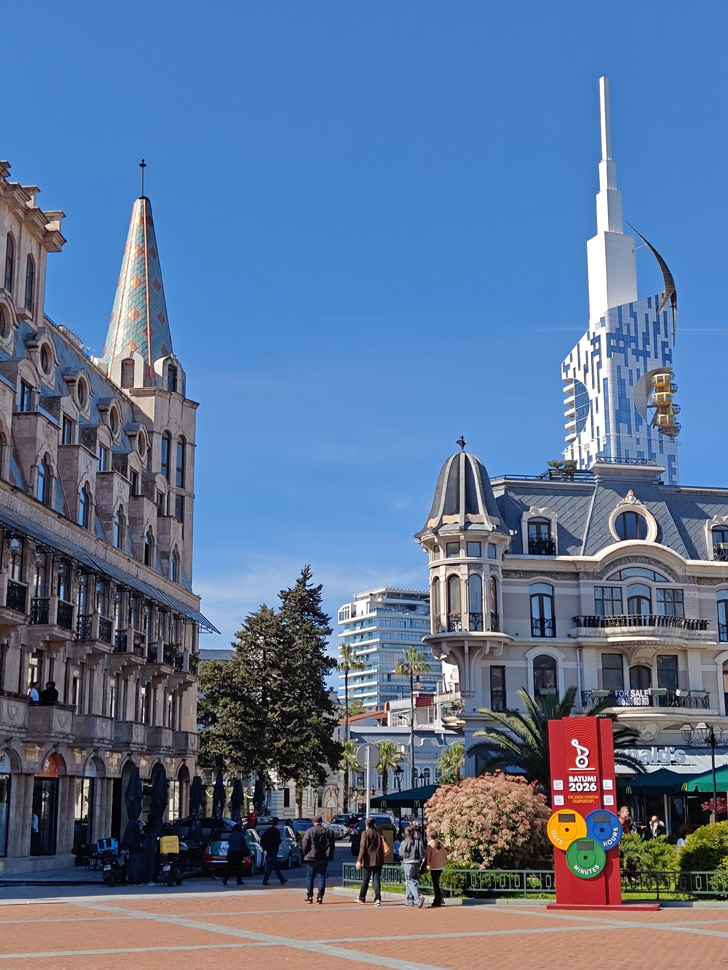 The image shows a sunny day in Batumi, Georgia, featuring distinctive architecture. On the left is a multi-story building with intricate stonework, arched windows, and balconies, topped with a pointed, tiled roof tower. To the right is a building with a domed turret, and behind it looms a tall, modern tower with a white and blue facade, with golden Ferris wheel cabins on the side.
In the foreground, a brick-paved plaza is visible with several people walking, some heading towards the camera. A red sign with the inscription "Batumi 2026" is planted at the plaza, along with the wording “DAYS”, “HOURS”, “MINUTES”. Greenery, including trees, shrubs, and palm trees, adds to the scenery. Several parked cars and scooters are visible in the background.