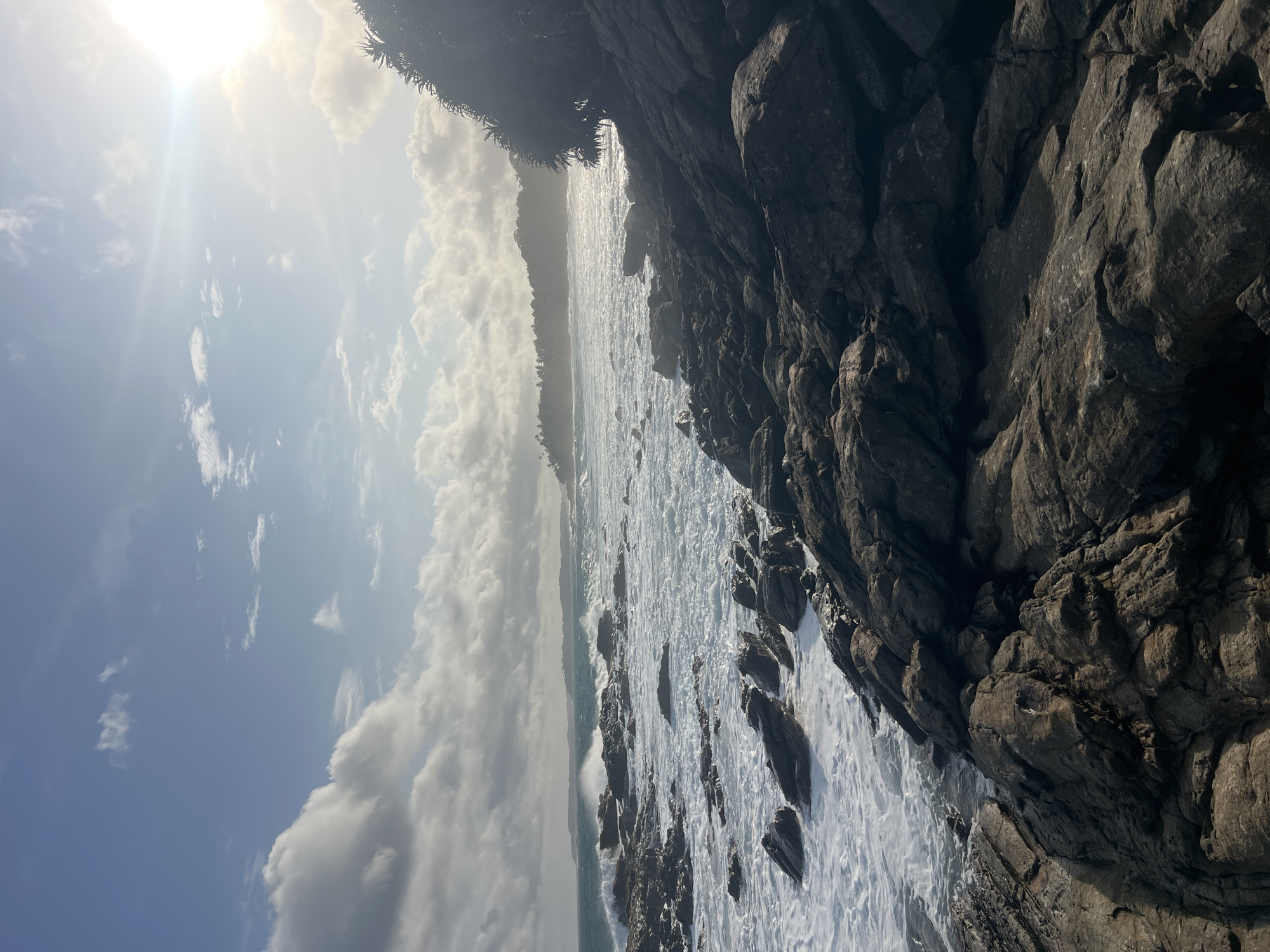 The image captures a rocky coastline in Tangalle, Sri Lanka, under a partially cloudy sky. The sun shines brightly from the upper right corner, casting a warm glow and creating lens flare. Waves crash against the dark, rugged rocks scattered across the foreground and midground, creating white foam and spray. The ocean extends to the horizon, where a distant, tree-covered shoreline is visible under a hazy sky. On the far right, lush green foliage frames the scene. There are no people or animals present in the image.