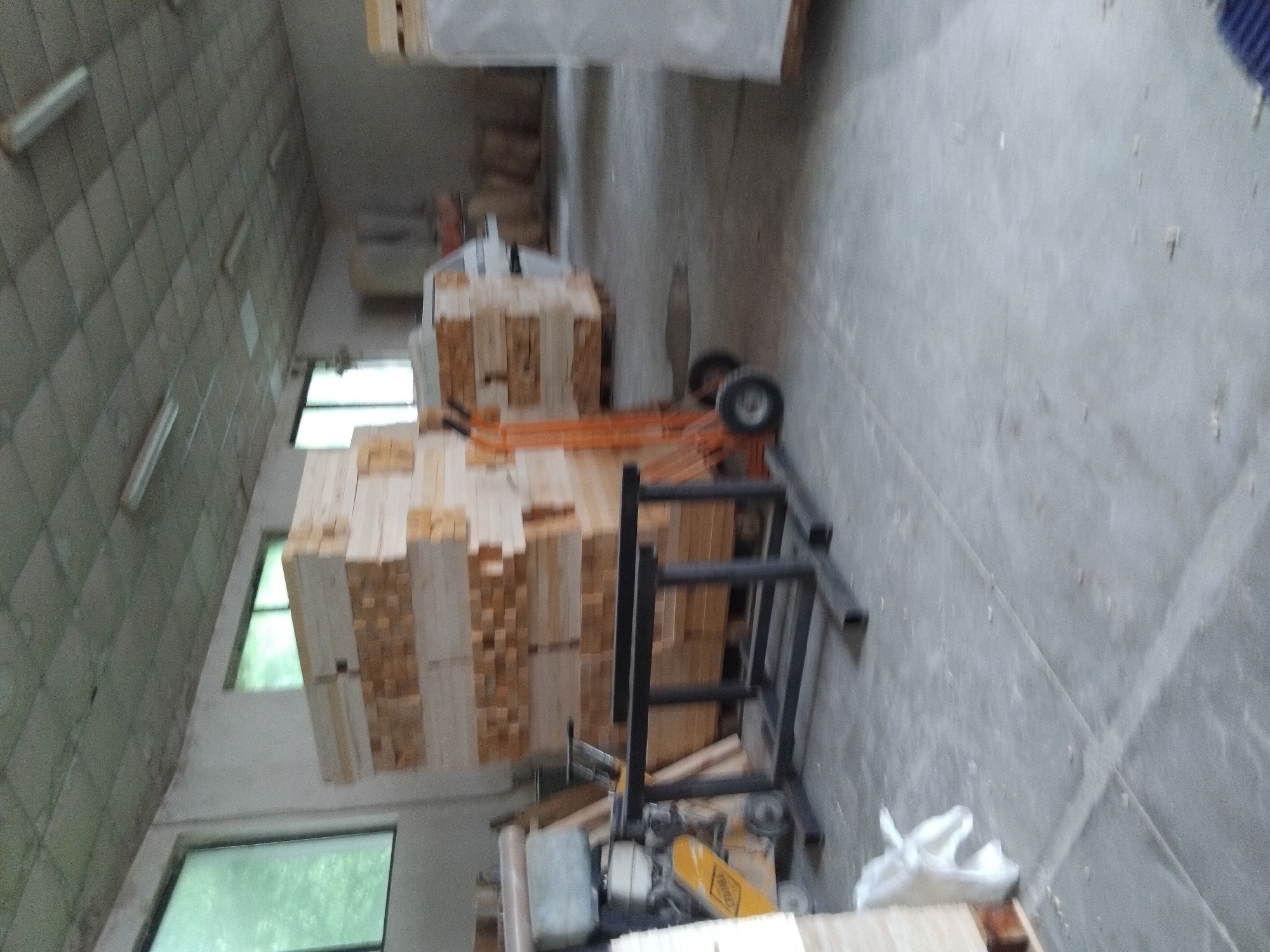 The image depicts the interior of a warehouse or storage facility in Unknown, Unknown. Stacks of lumber are visible, some strapped together in bundles and others appearing loose. A hand truck is positioned near one of the stacks, presumably used for moving the lumber. Various pieces of equipment, including what appears to be a saw, are also present in the scene. The environment suggests an industrial or storage space, with visible windows providing natural light.