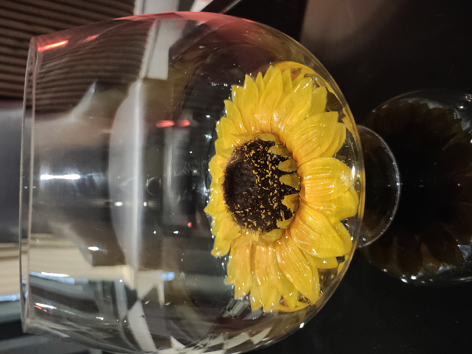 A close-up shot shows a single, vibrant yellow sunflower submerged in clear liquid within a large, transparent glass bowl. The sunflower is positioned centrally, its petals spread wide, with a dark brown, textured center. The liquid reflects light, creating shimmering highlights on the petals and the surface of the water. The glass bowl has a rounded shape, tapering to a stem and base that are also visible, creating a subtle reflection on the dark, glossy surface it rests upon. The background is indistinct, appearing as blurred shapes and colors, possibly suggesting an indoor setting with artificial lighting. The image does not contain any information that would allow for its location to be specified as Manikata, Malta.