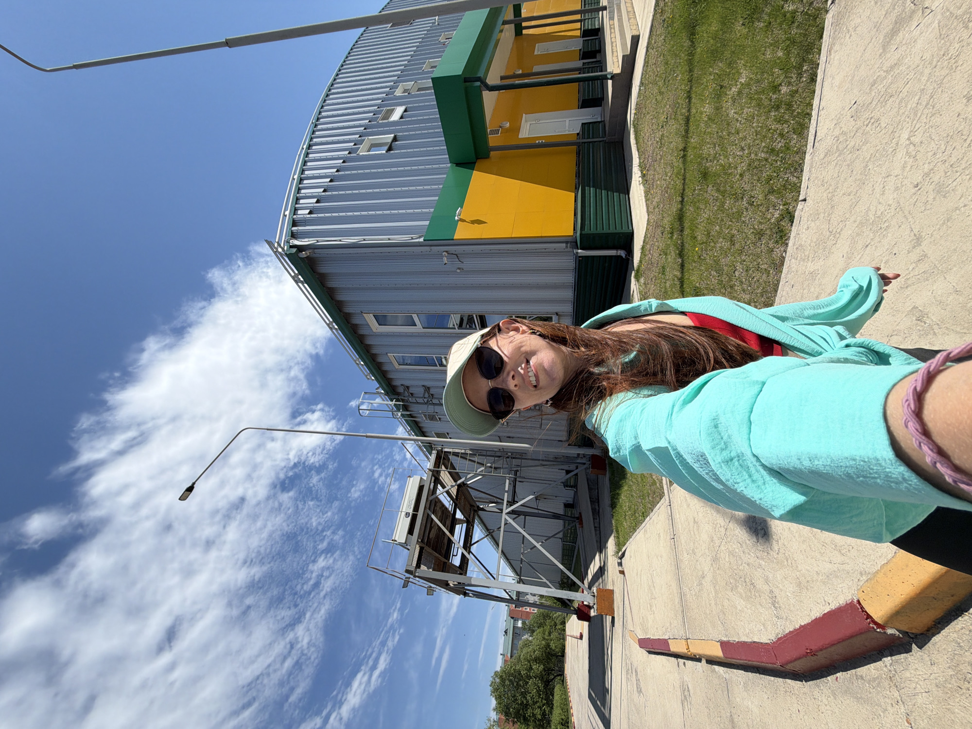 A woman in sunglasses and a hat takes a selfie outdoors, with a large, industrial-style building in the background. The building is primarily silver with a section painted yellow and green. A metal structure resembling a tower is also visible. The sky is blue with scattered clouds. The location is likely Birobidzhan, Russia. The woman is standing on a sidewalk with a painted curb.