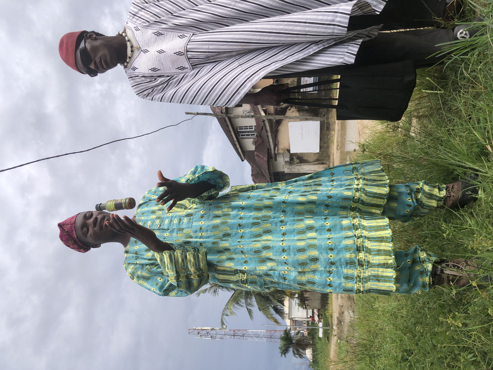 In the city of Ado, Nigeria, two men stand outdoors in a grassy area, possibly near a village. The man on the left is wearing a traditional blue and green patterned robe, a red patterned headscarf, and brown boots. He is performing a trick, with a small bottle of liquid hovering in the air between his hands, which are outstretched as if to catch it. The man on the right is dressed in a white and black striped robe, dark pants, and black sunglasses. He wears a necklace of black and white beads and is carrying a black tote bag. In the background, a cell tower and a two-story building are visible. The sky is overcast.