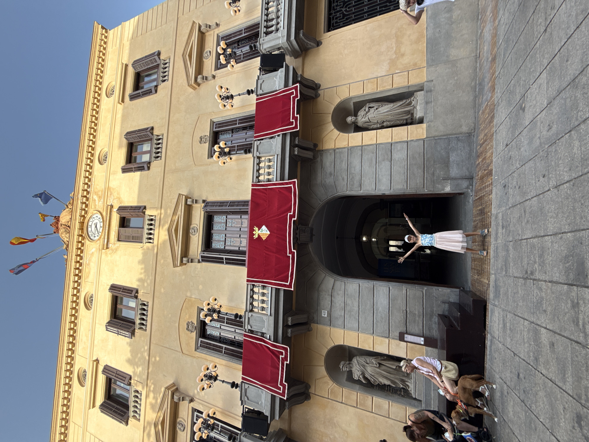 The image shows a wide, low-angle view of the exterior of a light yellow building, likely a public building in Mataró, Spain, given the crest displayed and flags. The building has a classical architectural style with multiple stories, visible windows with dark brown shutters, and balconies adorned with red velvet drapes bearing a coat of arms. Several ornate lamps with multiple bulbs are mounted on the balconies. At the top of the building, there's a decorative cornice, a clock face, and flags on poles.
In the foreground, a young person in a light-colored dress and a white cap stands with arms outstretched, facing the building, appearing to pose for a photograph. To the left, a few people are seated on a raised platform or steps. One woman wears a dark tank top and shorts, another woman wears a white tank top and a beige skirt, and a brown dog is near them. The ground in the foreground is paved with large, grey stone slabs. An arched entryway is visible in the center of the building, leading into a darker interior. The overall scene is set outdoors under a clear blue sky.