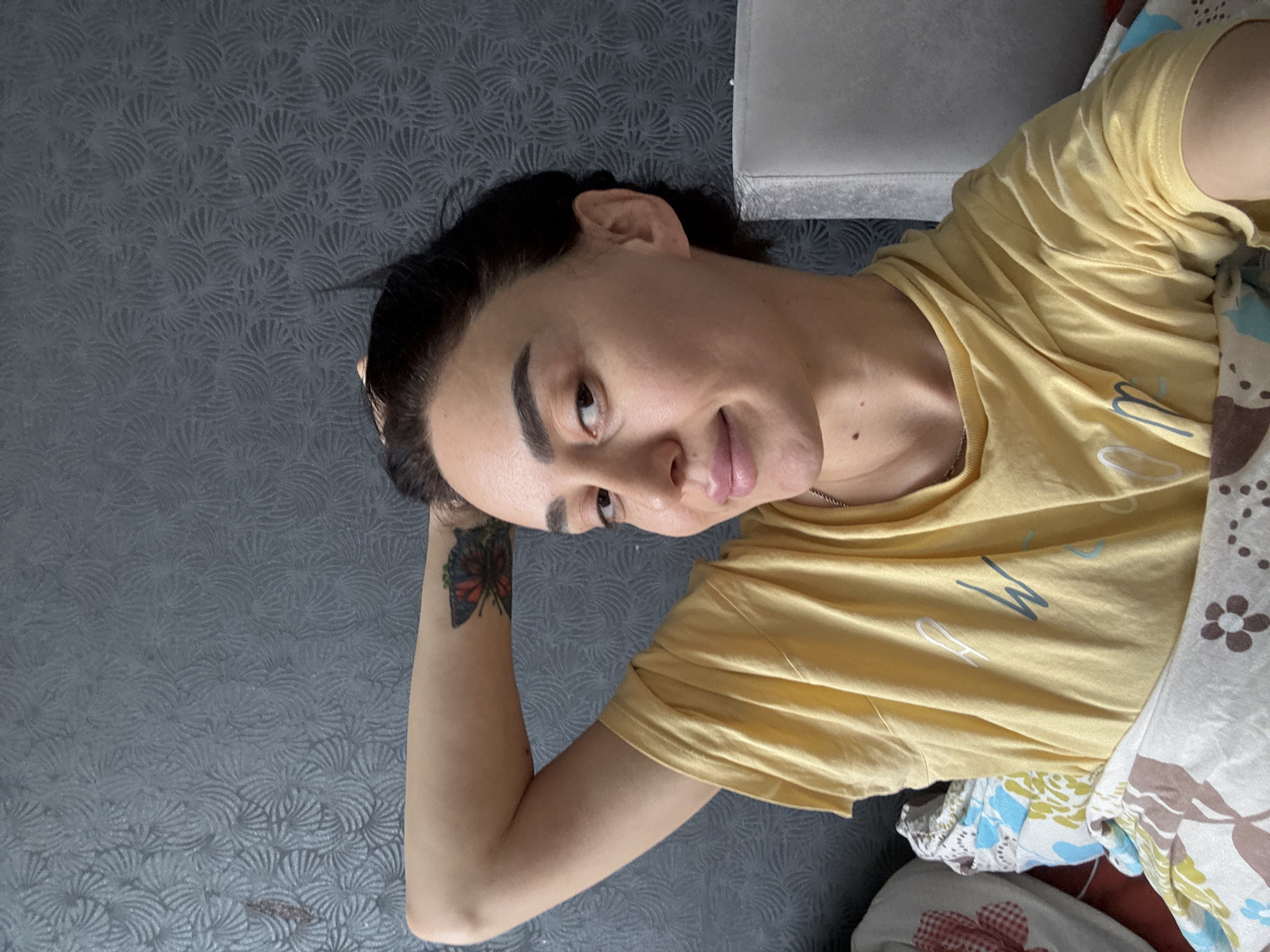 The main image shows a person, likely female, with dark hair, lying down and looking towards the camera with a gentle smile. The image is rotated 90 degrees clockwise, positioning the person's head on the right side of the frame and their body extending towards the left.
The individual wears a light yellow t-shirt. Visible on the t-shirt are some light blue and white letters or designs, including a clear "M" in blue. A thin, silver-colored chain is worn around their neck. A colorful butterfly tattoo, featuring red, black, and blue, is visible on their left forearm. Their left arm is bent, with the hand resting near their head.
The background consists of a dark grey wall or headboard featuring a distinct textured pattern of swirling or fan-like motifs. A light grey object, possibly a cushion or part of a bed, is visible behind the person's head. The lower part of the frame shows patterned bedding or a blanket in shades of blue, white, brown, and hints of red, partially covering the person's body. The setting appears to be an indoor space, likely a bedroom or living area. The lighting suggests it is daytime. The person's expression is relaxed and comfortable. No other specific text is clearly discernible. The location context provided is Tbilisi, Georgia.