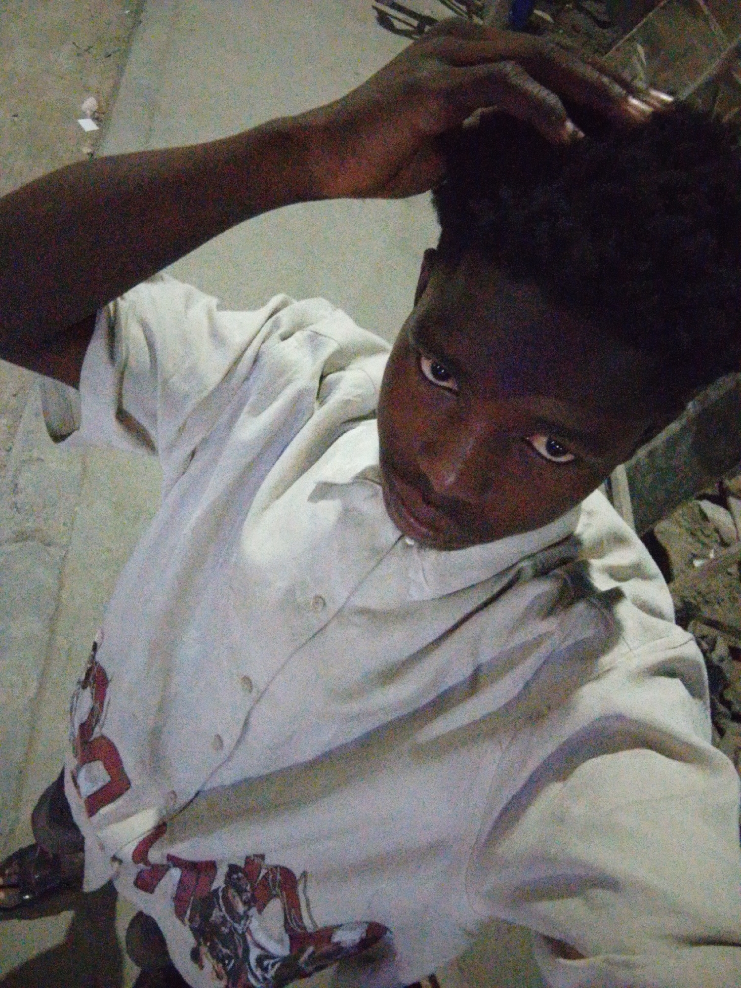 A person with dark skin and short, curly black hair is standing on a concrete surface. He is wearing a light-colored button-down shirt with a graphic design on the lower front. The person is looking up at the camera with a hand on their head. The background is dimly lit, suggesting an outdoor setting. Based on the prompt, the location is the city of Lagos, Nigeria. There are hints of construction debris or rubble visible on the right side of the frame.