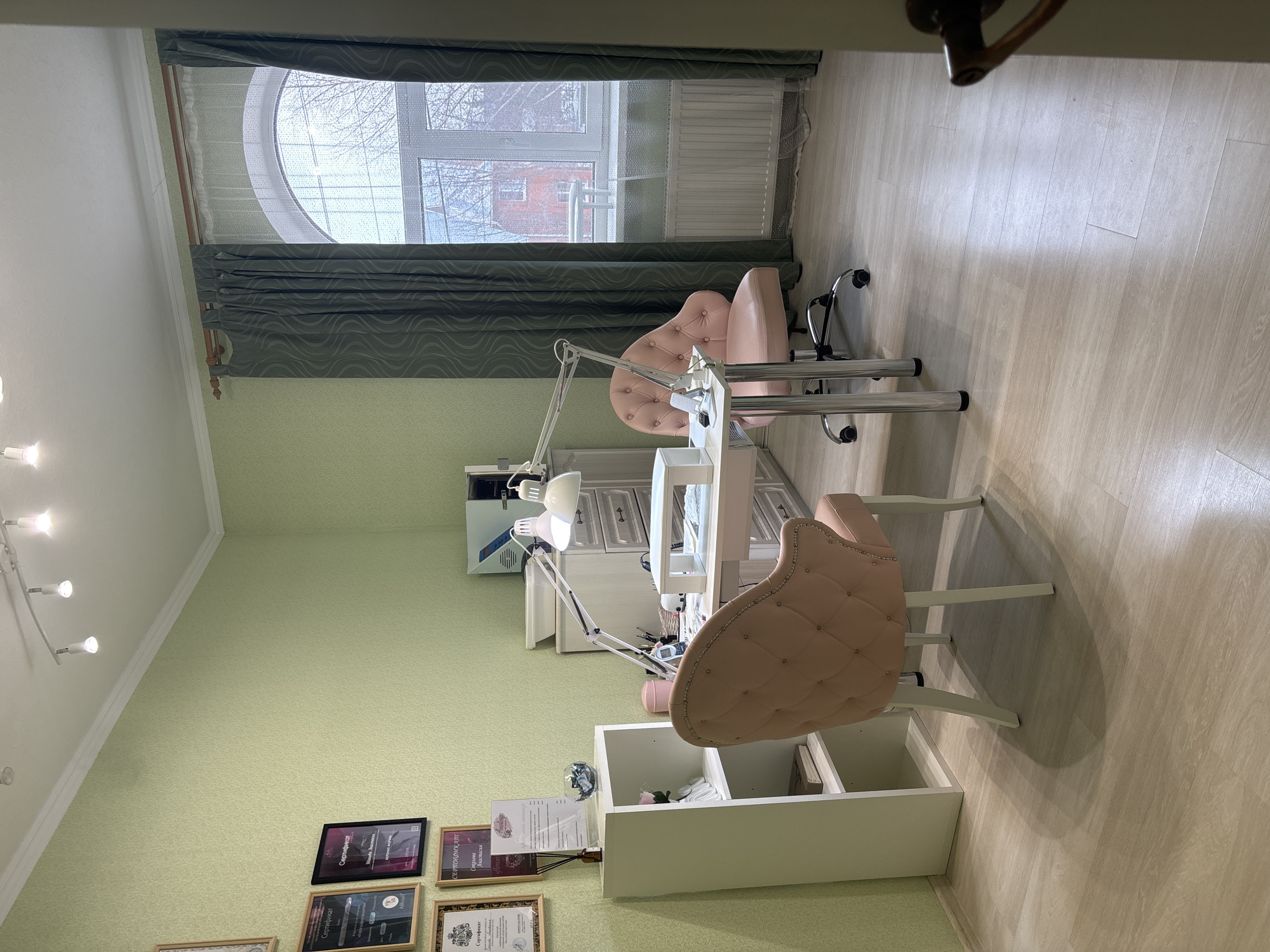 The image depicts an unoccupied interior room, functioning as a nail salon or beauty treatment area, located in Shul'gino, Russia. The walls are covered in light green, subtly patterned wallpaper. The flooring consists of light-colored wood-effect laminate panels. The white ceiling features a three-spotlight track lighting fixture.
On the right side of the room, a large, arched window is fitted with dark green, wave-patterned curtains and a sheer white curtain. Outside the window, bare tree branches and a distant red building are visible, suggesting an overcast or winter day. A white radiator is installed beneath the window.
Against the left wall, a white open-shelf unit displays a pink artificial flower, a reed diffuser, a glass bowl containing small items, and a book on the lower shelf. Above and to the left of this unit, multiple framed certificates or diplomas are affixed to the wall.
In the center of the room, a white manicure table with integrated drawers serves as the primary workspace. A white, dual-headed adjustable desk lamp is clamped to the table. On the table, a white nail dust collector with a hand rest, a smartphone in a stand, and various manicure tools and products are arranged. Two pink, tufted armchairs are positioned at the table: one stationary chair facing the table from the front, and one rolling, five-wheeled office chair on the opposite side.
Behind the manicure table, against the back wall, a light-colored storage cabinet features a grey countertop. On this countertop, a light blue and white sterilization unit and a white tray are visible. No individuals are present in the scene, and no actions or interactions are depicted.