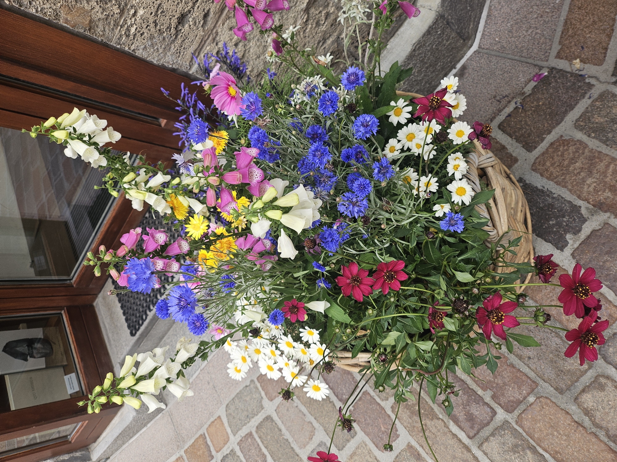 The image depicts a woven basket overflowing with a vibrant arrangement of wildflowers. The flowers are a mix of colors including white, pink, yellow, blue, and deep red. Specifically, visible are foxgloves, cornflowers, daisies, and what appear to be dahlias or cosmos. The basket sits on a cobblestone pathway in front of a wooden door with glass panels. The door is flanked by a textured stone wall on the right. In the background, visible through the glass door, is an interior space with what looks like display shelves. The setting is outdoors, suggesting it might be an entrance to a shop or establishment in St. Moritz, Switzerland. No people are present in the image. The lighting suggests it is daytime.