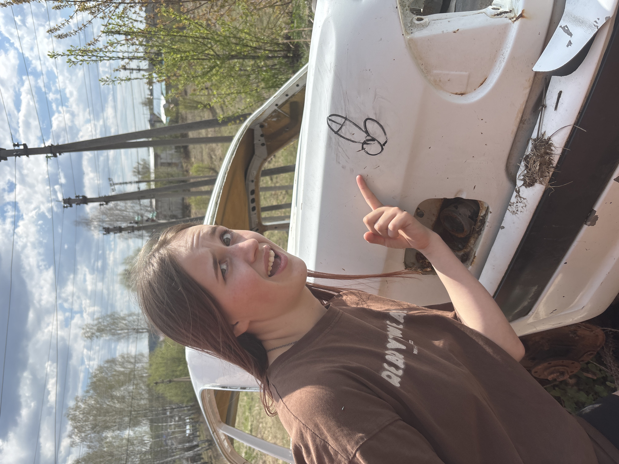 A young woman stands outdoors, pointing at a crude drawing of male genitalia on the side of a white, severely damaged car. The woman has long brown hair and wears a brown t-shirt. The car appears to be in a state of disrepair, with rust and debris visible. The background shows a rural setting with trees and utility poles. The sky is partly cloudy. The location may be in or near the city of Фоки, Russia, given the user's prompt.