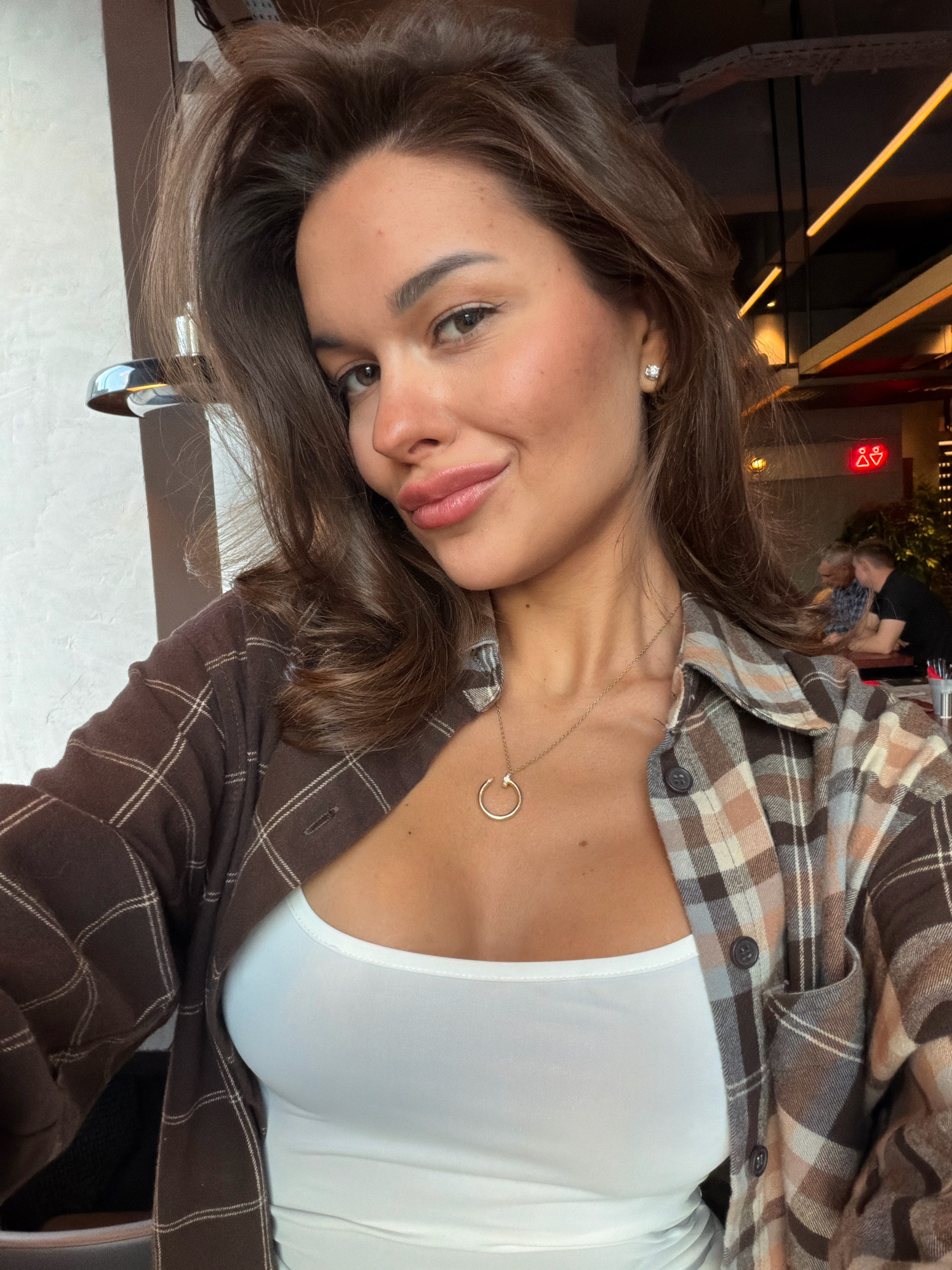 A selfie taken indoors of a young woman with brown hair, looking at the camera. She is wearing a white tank top and an open brown and beige plaid shirt. She has a gold necklace with a circular pendant. Her makeup is visible, with eyeliner, eyeshadow, and lipstick. In the background, blurred figures of people can be seen at tables in what appears to be a restaurant or cafe, with ceiling lights and decorations. The overall impression is a casual portrait within an urban public setting.