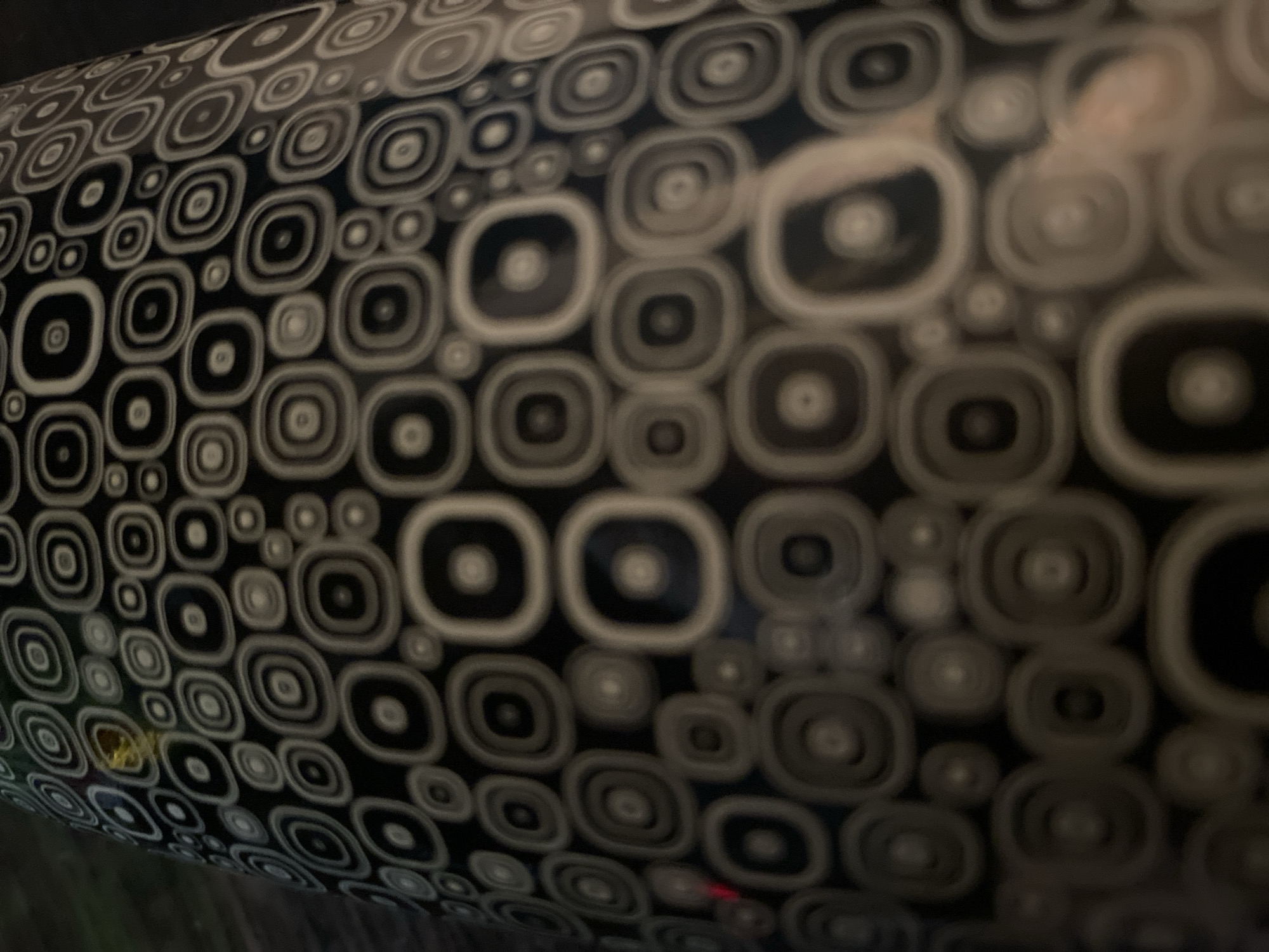 The image shows a close-up of a cylindrical object with a repeating pattern of concentric circles and rounded squares. The pattern alternates between black and light gray/white shapes. The object appears to be a vase or a decorative container. The lighting is dim, casting shadows and creating a blurred effect. There is a small area of yellow discoloration on the upper left portion of the object. The background is dark and out of focus, suggesting it is an indoor environment. There is no discernible information about the city of Ivanteyevka, Russia, or any other specific location, people, actions, or interactions within the image.