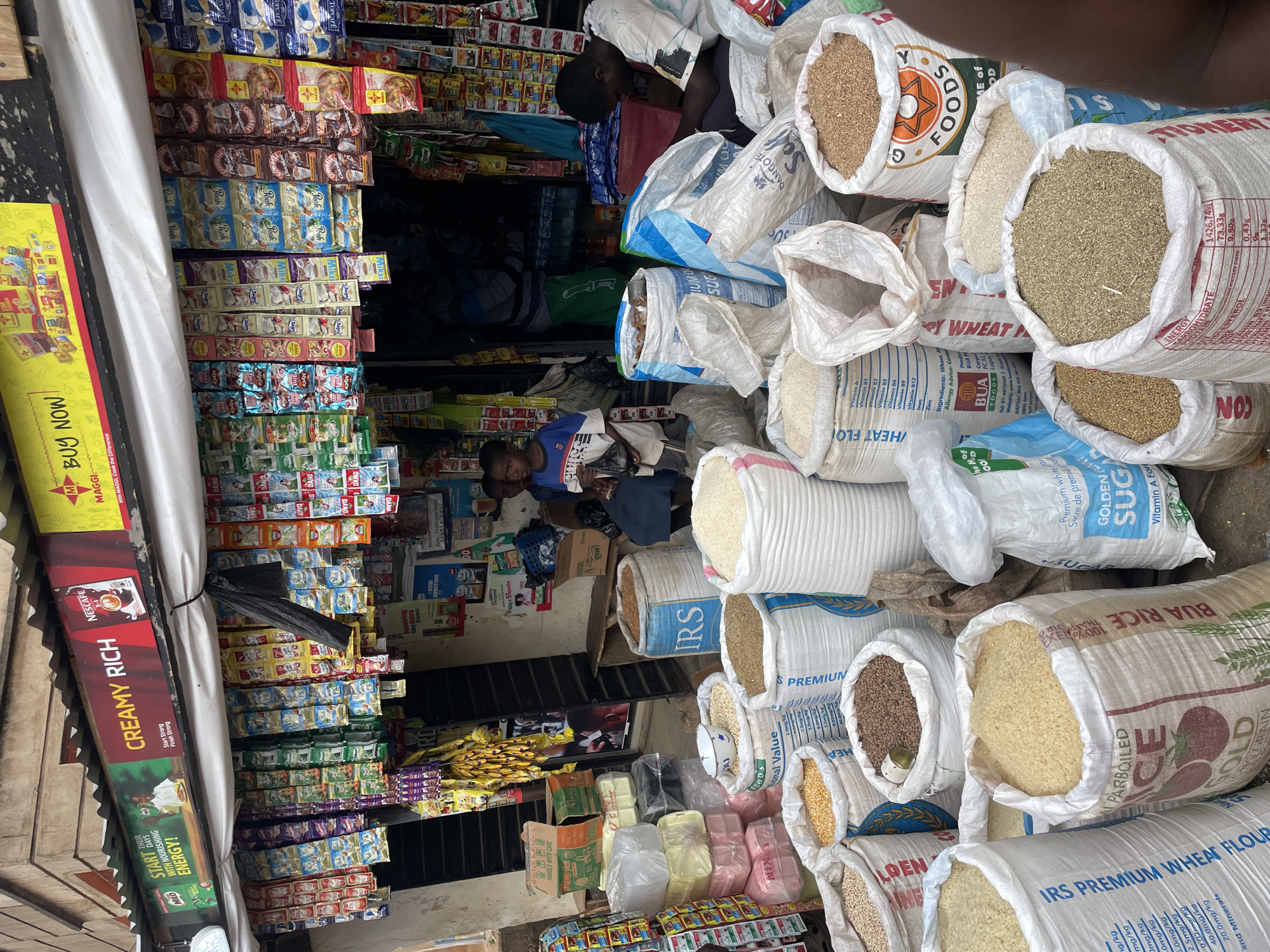 This is a detailed, eye-level photograph of the interior and exterior of a small retail shop or stall, likely in Jos, Nigeria, filled with a variety of food items and consumer goods.
Several large, open bags filled with grains (rice, wheat, and possibly other milled products) dominate the right side of the frame. The bags display brands like “IRS” and “BUA RICE.” Packages of smaller grocery items fill the shop. They sit on shelves and on the floor. There are numerous packages of powdered milk, cocoa and other food products. The shelves are stacked with various brands.
In the interior, at least two individuals are visible. One person appears to be lying down or resting amongst the bags of grains, while another sits further inside the store, seemingly engaged in conversation or reading.
Advertising and promotional materials line the exterior and the shelves inside, with brands like "NESCAFE," "MILO," "MAGGI," and "DANO" prominently displayed. The overall scene depicts a busy retail environment catering to the local population.