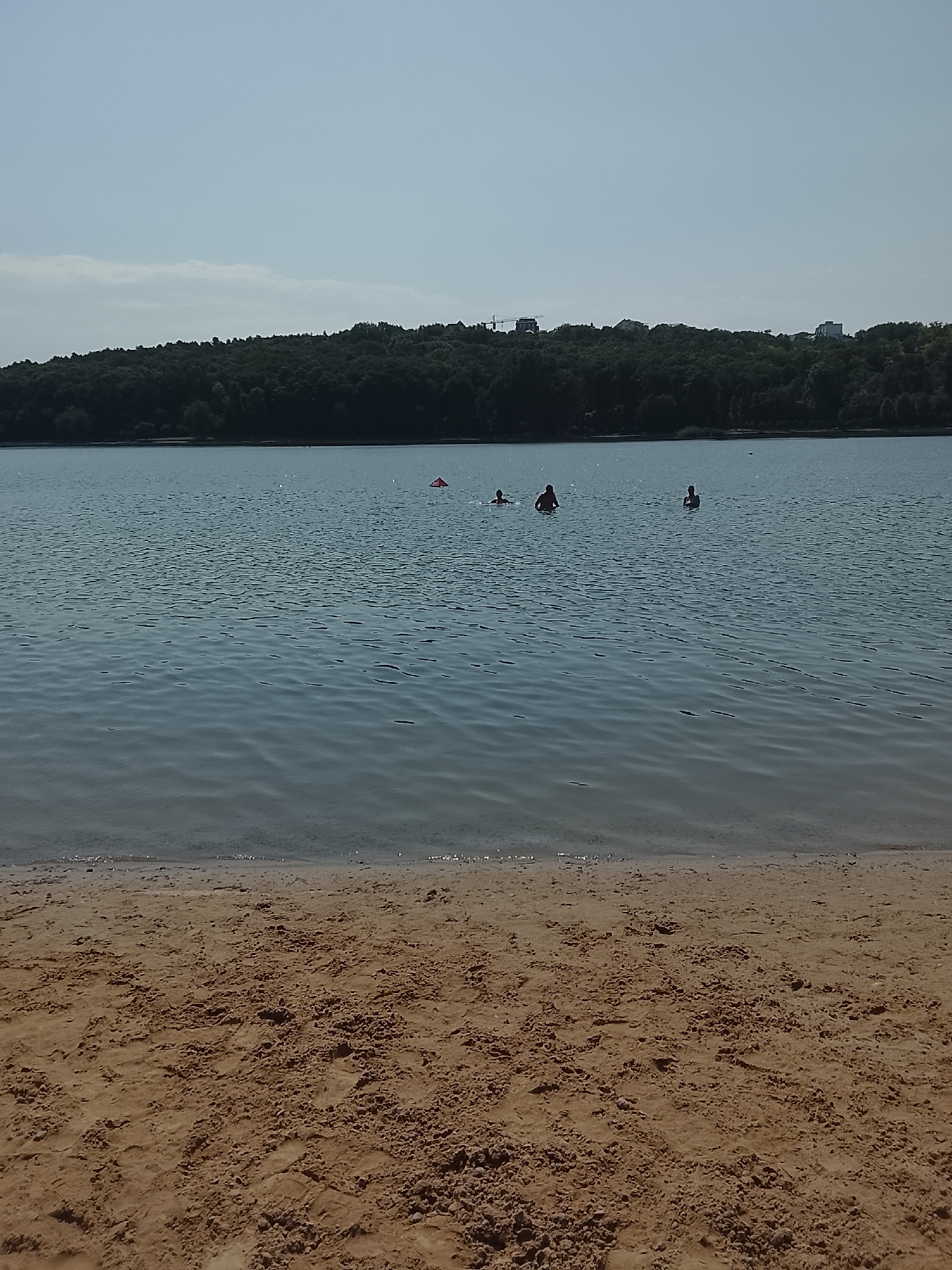 The image depicts a serene scene at a lake on a clear day. In the foreground, a sandy beach stretches out, its texture detailed with small bumps and depressions. The water of the lake is calm, with subtle ripples reflecting the light blue sky.
Further out on the lake, several people are enjoying a swim. Three individuals are visible as dark silhouettes against the water. One person is further to the left, with two others closer to the center. A bright red buouy floats in the water to the right of the swimmers.
The background is dominated by a dense, dark green forest that lines the far shore of the lake. Beyond the trees, a few distant buildings and a construction crane are visible, suggesting the proximity of an urban area. The overall impression is one of a tranquil natural escape within or near a city.
This scene is consistent with the Văduleni Lake, a popular recreational area located on the outskirts of Chișinău, Moldova. The lake is known for its sandy beaches and forested surroundings, offering a place for swimming and relaxation for residents of the capital city.