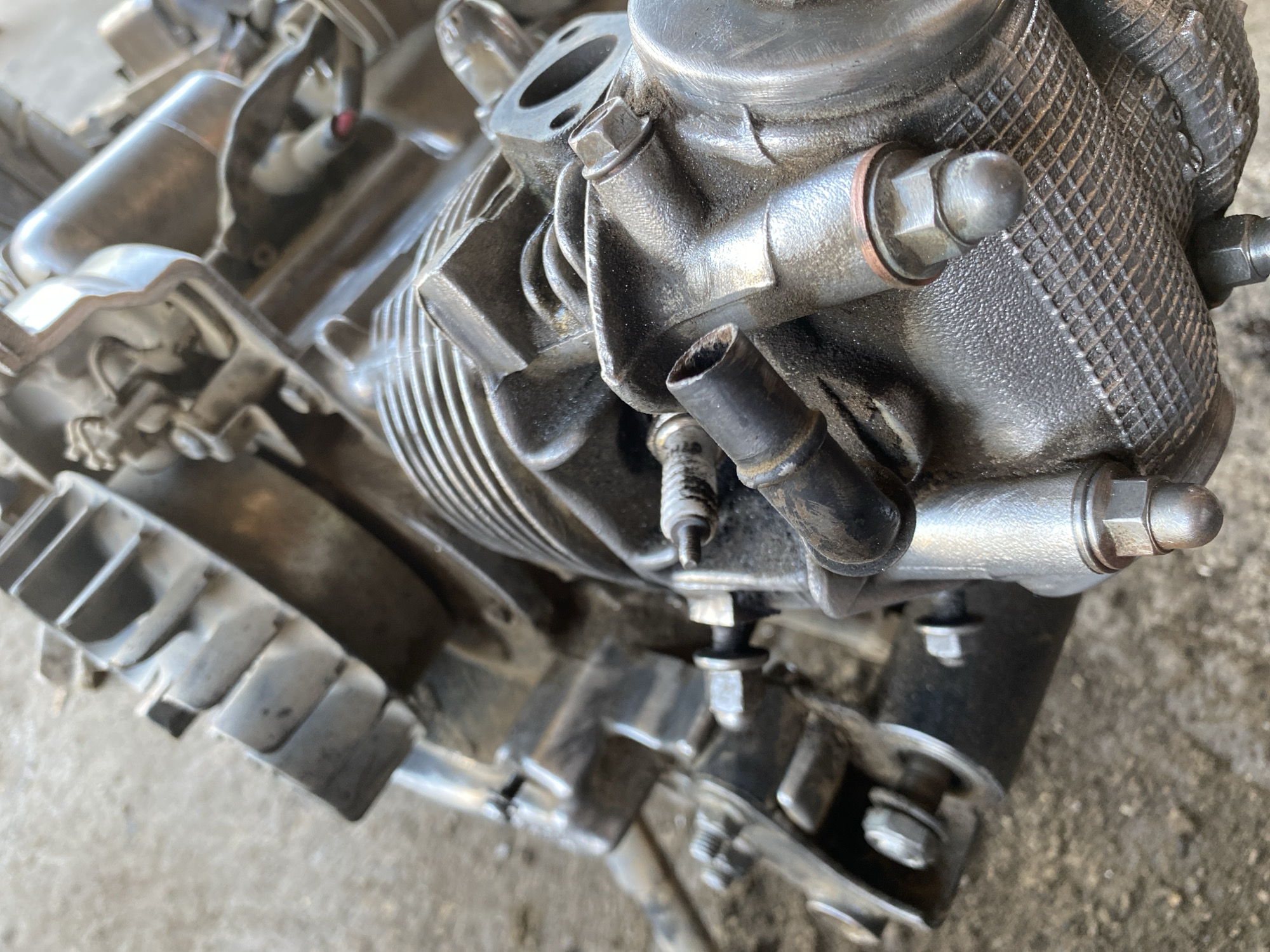 A close-up, eye-level, outdoor, slightly low-angle, medium shot captures a detailed view of a heavily soiled, metal engine part, possibly from a motorcycle or small vehicle, resting on a concrete or asphalt surface. The engine component is predominantly silver and grey, with a ribbed texture on one side and a diamond-patterned texture on another. Various bolts, nuts, and tubes are visible, some appearing slightly rusted or worn. One notable feature is a black tube protruding from the engine. The background is blurred, suggesting a shallow depth of field, and shows the rough texture of the ground. The image lacks any human presence or discernible signs of a specific location within the city of Bayu, Indonesia; it is a generic depiction of mechanical debris.