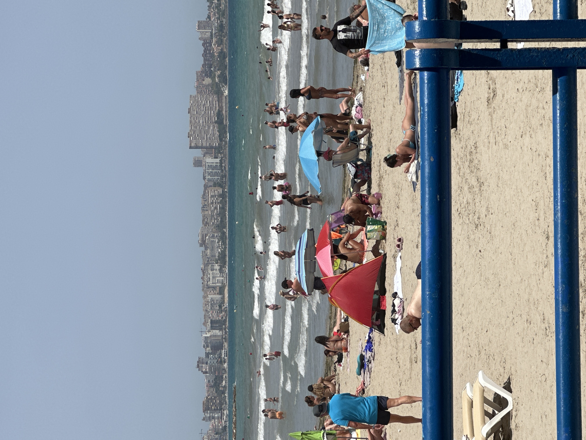 This is a photograph of a crowded beach on a sunny day, with a city skyline in the background. The image is oriented vertically. People are engaged in various beach activities, including swimming in the ocean, sunbathing on the sand, and relaxing under umbrellas. The sea is choppy with visible waves, and there are many people in the water. Several colorful umbrellas and beach chairs are scattered across the sand. The city skyline features numerous tall buildings, suggesting an urban coastal setting. The time of day appears to be midday, given the bright sunlight and shadows cast. The overall atmosphere is one of leisure and recreation. No visible text is present in the image.