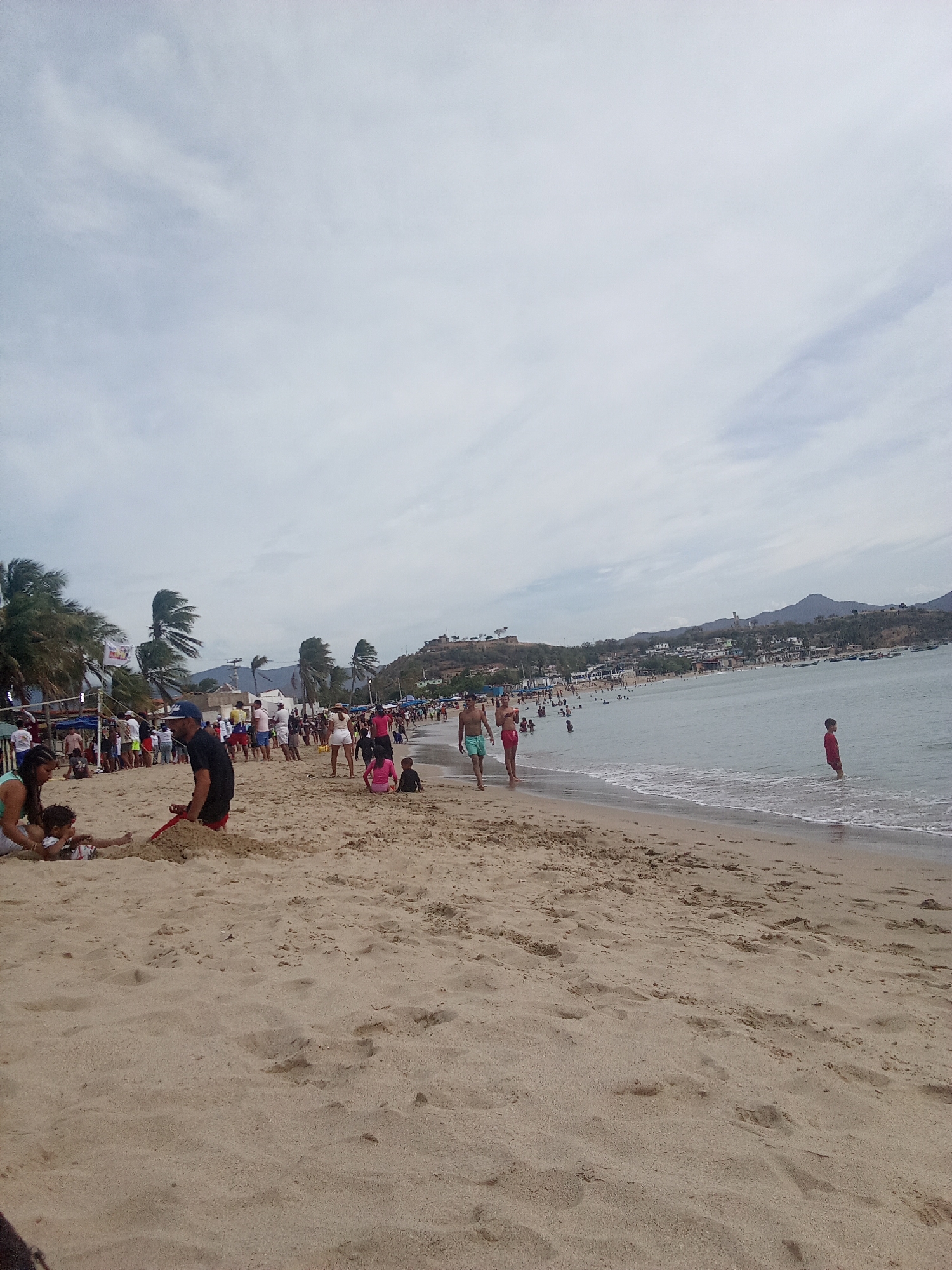 The image depicts a crowded beach in Juan Griego, Venezuela. The beach is sandy with many footprints and impressions. There are numerous people present, including adults and children. Some are interacting on the beach, such as digging in the sand, while others are wading in the water. Palm trees line the edge of the beach. In the background is a town with buildings and hills. The sky is overcast.