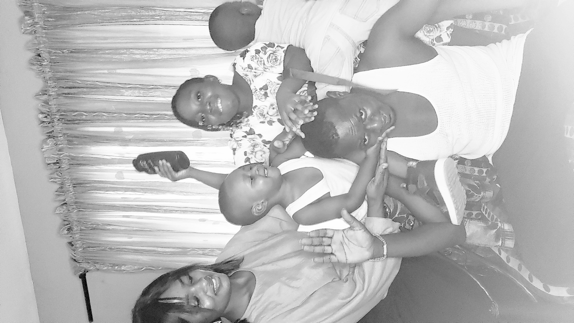 The image captures a group of people posing together indoors. The setting appears to be a home, as evidenced by the curtain in the background. Four people are visible, including what looks like a mother or older sister and three children. One of the children is holding a small, rectangular object that appears to be a camera or phone. They are interacting with each other, laughing, and looking towards the camera. The scene appears playful and familial. The location details are not discernible from the image.