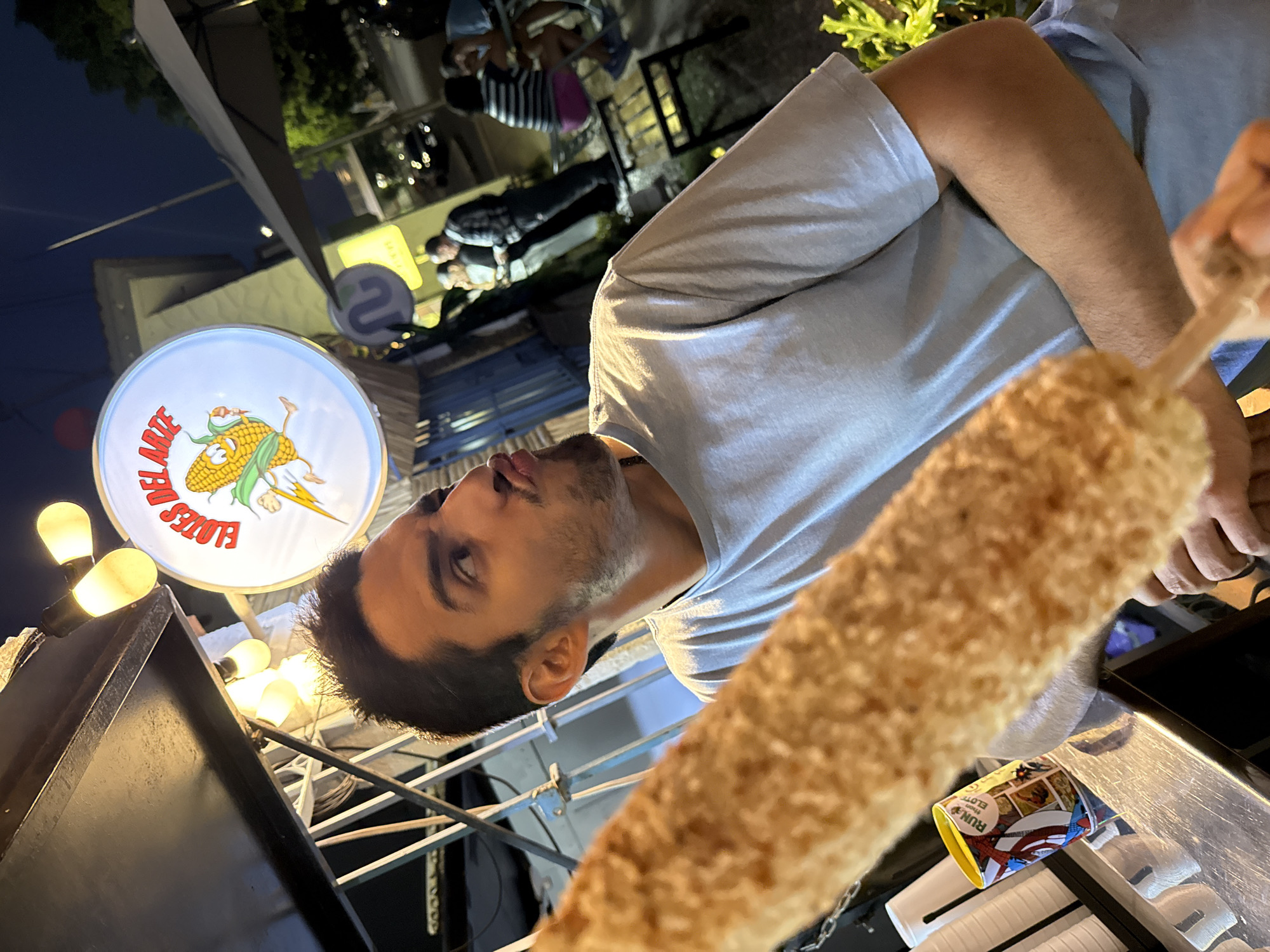 A man with a light blue shirt is holding a corndog, with a sign for Elotes Del Arte visible in the background. Other people can be seen behind a light-colored awning. The presence of the Elotes Del Arte sign indicates that this photograph was taken in Puebla, Mexico.