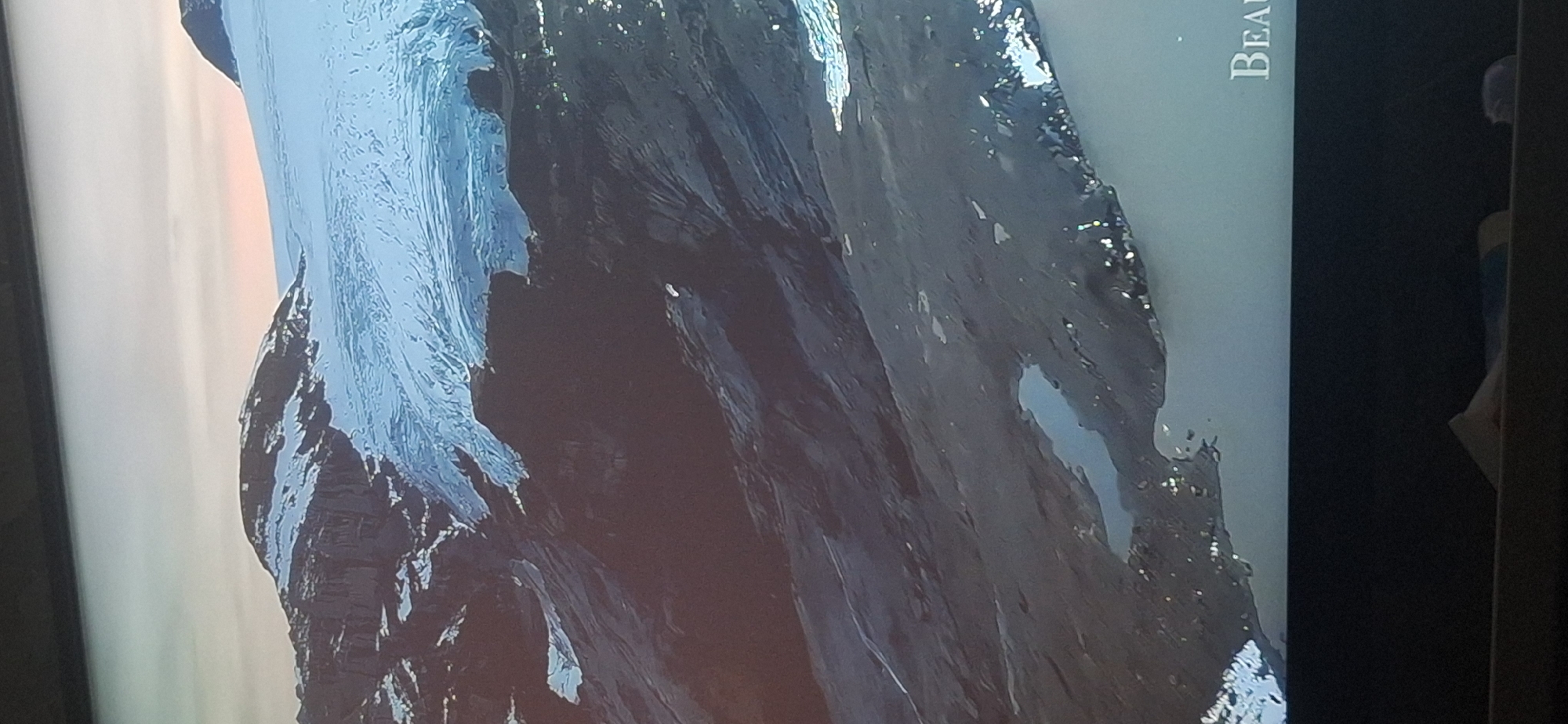 The image shows an aerial view of a vast glacier covering mountainous terrain. The glacier is a brilliant white and light blue, with visible crevasses and undulating surfaces. Dark, rocky mountains with patches of snow rise from the edges of the glacier. In the foreground, the landscape transitions to dark, rugged terrain with several small, still lakes reflecting the sky. The sky above is a soft gradient of pale yellow and pink, suggesting either sunrise or sunset. In the bottom right corner, the word "BEAU" is visible in large, serif font, possibly part of a larger title or watermark. The image appears to be a photograph or screenshot, possibly of a documentary or natural landscape presentation. There are no people or specific city landmarks visible that would indicate a location in Bila Tserkva, Ukraine.