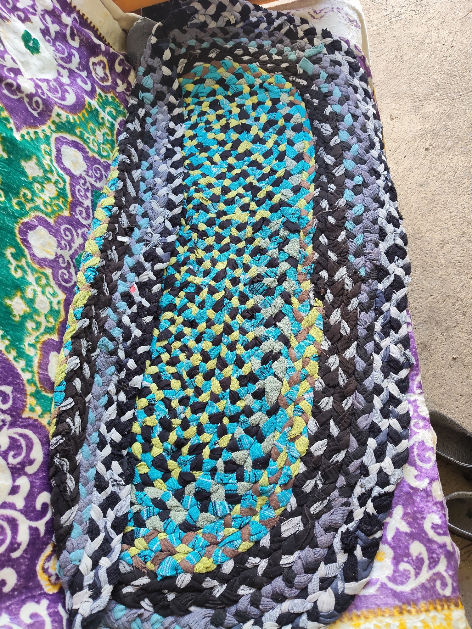 The image shows a braided rag rug with an oval center of blue, yellow, and black woven strips, bordered by concentric bands of black, grey, blue, and white braided fabric. The rug is laid on top of a larger fabric with a purple and green paisley-like pattern. The scene is outdoors, with a concrete surface visible to the right of the rug. The overall impression is one of a rustic or homemade item, possibly displayed or stored outside. The location is identified as Mîndîc, Moldova.