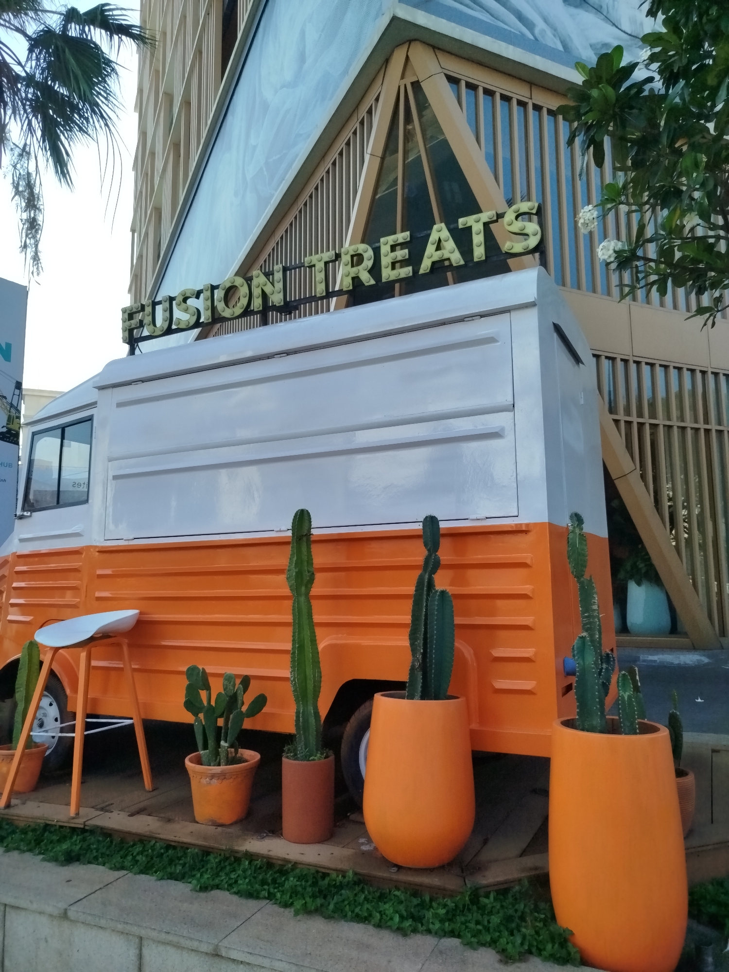 The scene shows a food truck named "Fusion Treats" in what appears to be Ho Chi Minh City, Vietnam. The truck is painted white and orange and has a line of potted cacti in front of it. A modern-looking building with a triangular design is visible in the background. A palm tree is visible in the top left corner of the image. A small, minimalist white stool is positioned near one side of the truck. The location appears to be an outdoor urban setting, with some greenery visible near the base of the image.