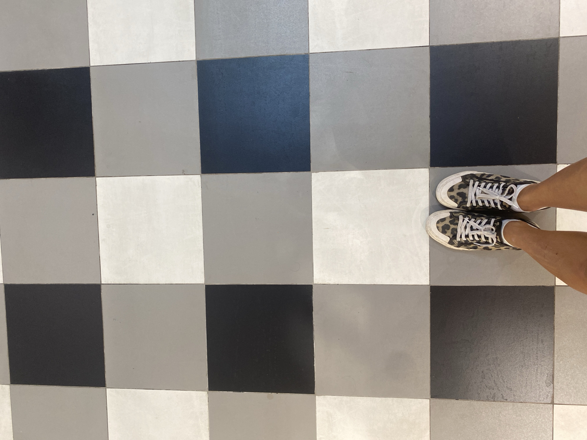 The image shows a person's feet wearing leopard-print sneakers standing on a checkered tile floor. The tiles are in shades of black, white, and grey, arranged in a checkerboard pattern. The scene appears to be indoors. There is no indication of the location in Denpasar, Indonesia.