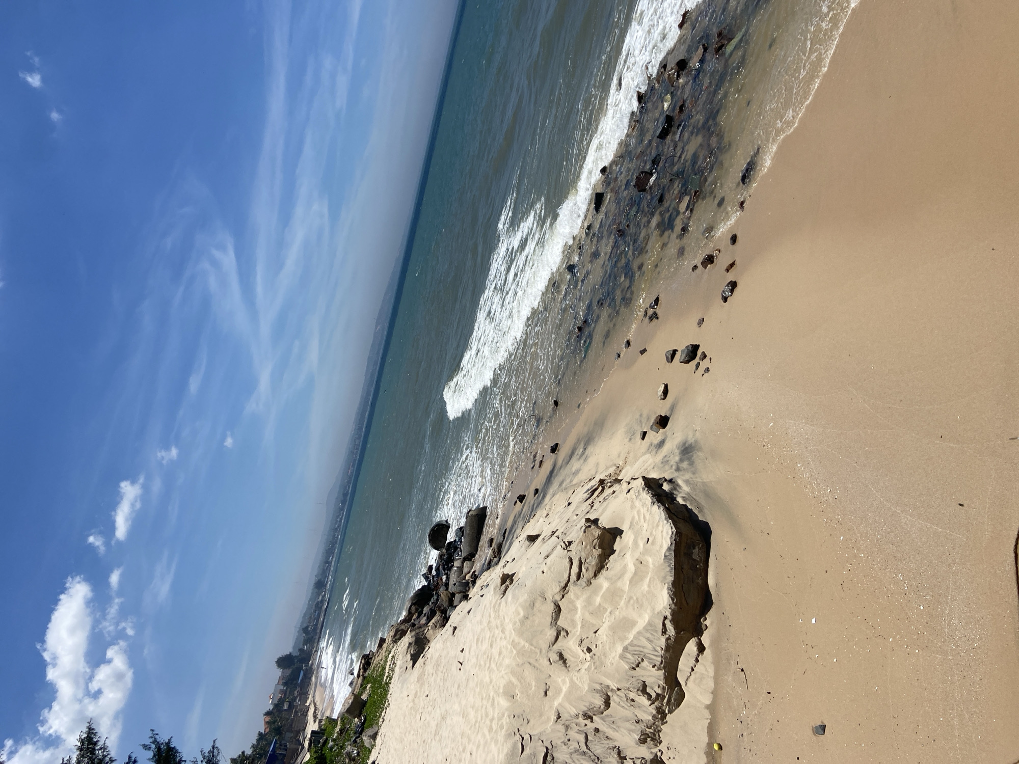 A wide-angle, eye-level view captures a scenic coastal environment in Phu Thuy Ward, Vietnam. The foreground features a sandy beach with dark streaks of wet sand, leading to the ocean. Jagged rocks and small pebbles are scattered along the shoreline. The sea is a deep blue-green, with gentle waves breaking near the shore. In the distance, a hazy shoreline is visible with a line of trees and some buildings. The sky is a vibrant blue, dotted with scattered white clouds. A lone figure is visible walking on the beach in the distance.
