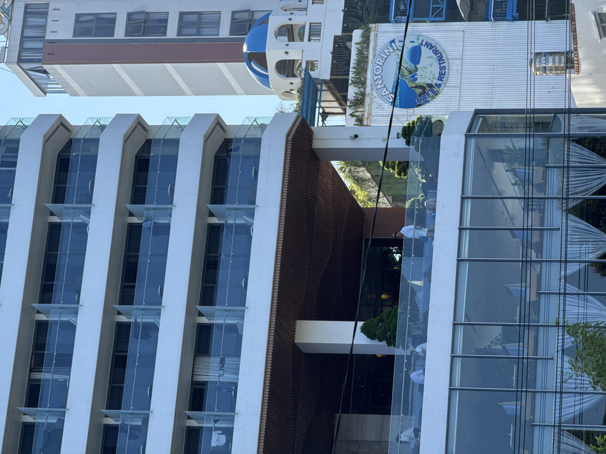 The image captures an urban scene in Nha Trang, Vietnam, focusing on two prominent buildings under a clear blue sky. The primary structure on the left is a multi-story white building, likely a hotel or residential complex, featuring numerous balconies with clear glass railings across its upper floors. Some balconies display items, including articles of clothing drying, and individual white chairs and small tables are visible on several.
Below the main block of balconies, an expansive open-air terrace is situated, supported by white columns and shaded by a dark brown, slatted ceiling. Approximately six individuals are present on this terrace. One person, dressed in a white uniform or robe, walks towards the right. Two other individuals in light-colored shirts are seated at a table on the left side of the terrace. Green potted plants or small trees are interspersed across the terrace area. A security camera is affixed to a white column on the terrace level. The floor below the terrace exhibits large, full-height glass windows revealing an interior space furnished with multiple chairs featuring light blue backrests. White drapes are visible behind the windows on the lowest captured level.
To the right, a taller, narrower building is partially visible. This structure displays sections of white, grey, and reddish-brown exterior finishes, with multiple windows, some of which are partially open. A distinctive blue-domed architectural feature, accompanied by white arched openings, is present on an upper section of this building, evoking Santorini island aesthetics. Below this dome, a large circular blue and white sign for "SANTORINI CAFE & RESTAURANT" is prominently displayed on a white corrugated facade. A stainless steel water tank, labeled "SON," is situated on a lower rooftop section of this restaurant building. Multiple black electrical wires traverse horizontally across the mid-ground of the scene. Green foliage is visible in the lower foreground and between the buildings.