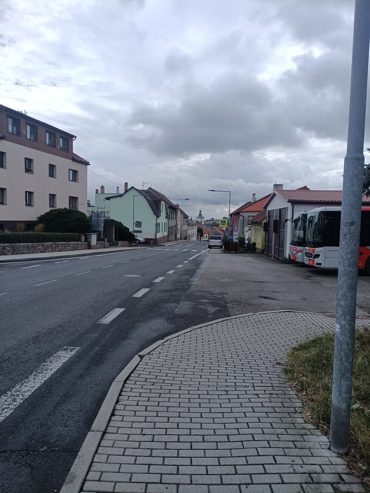 The image captures a street scene in Dalovy, Czechia, on an overcast day. The perspective is from a sidewalk looking down a road lined with buildings. A prominent light pole stands to the right. On the left side of the street, a multi-story building with a light-colored facade and a reddish-brown roof occupies the foreground. Further down, a row of buildings with varying colors, including pale green and muted tones, continues along the street. On the right side, a bus depot or parking area is visible, with two buses parked. One bus is white with red accents, and the other is predominantly red. The road has white lane markings, and there's a paved sidewalk with a brick pattern in the foreground. A few vehicles, including a white car and a smaller silver car, are present further down the road. In the distance, a church steeple is visible above the buildings. The sky is heavily clouded, contributing to the muted lighting of the scene.