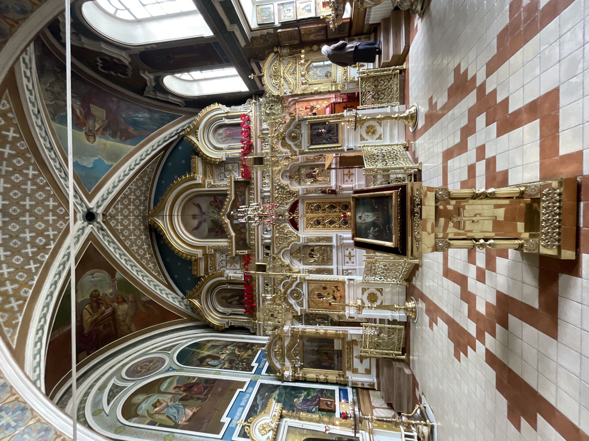 The image shows the interior of a Russian Orthodox church in Kazan, Russia. It features an ornate, gold-and-white iconostasis, or screen of icons, positioned in front of the altar. Paintings adorn the walls and the vaulted ceiling, depicting religious figures and scenes. The floor is tiled with a distinctive red and white geometric pattern. At the top of the image, natural light streams in through arched windows. One person is partially visible in the upper-right corner of the image.