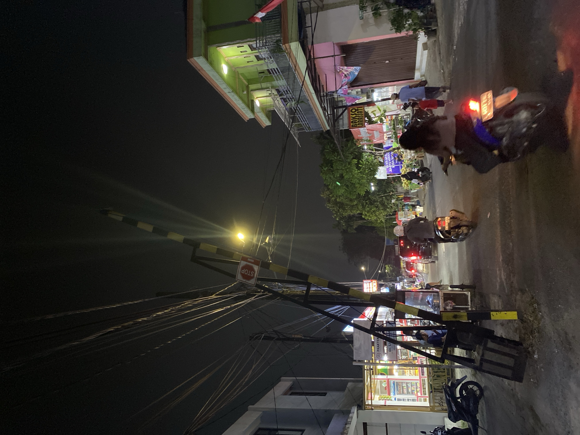The image captures a street scene at night in Tangerang, Indonesia. A prominent railway crossing gate, painted with yellow and black stripes, dominates the foreground and is partially lowered, suggesting an approaching train or recent passage. Overhead, a dense network of electrical wires crisscrosses the sky, illuminated by a streetlamp and ambient lights from the buildings.
On the right side of the street, a two-story building with a bright green facade stands, featuring balconies and windows. Below, a pink-fronted structure has a closed wooden door and several signs, including one for "BAKSO LARASSATI". A red and white Indonesian flag is visible on the upper floor.
On the left side, another building has a lighter colored facade and windows, with some signage visible, including a sign for a "PRAKTEK" (practice or clinic). A pharmacy appears to be open with its interior lights on, displaying shelves of products.
The street is active with various forms of transportation. Several motorbikes are present, some parked and others being ridden. One motorbike in the foreground is ridden by a woman wearing a light-colored shirt and dark pants, her face obscured. Another motorbike further down the street is ridden by a person wearing a helmet. Cars and other vehicles are also seen in the distance, indicating traffic.
Several people are visible: a man and a woman walking on the right side of the street, a child running between them, and individuals near the railway crossing gate and the pharmacy. The overall atmosphere is that of a bustling urban environment at night, with the sounds of traffic and the glow of lights from shops and buildings. The ground is unpaved or roughly paved, typical of some streets in the region.