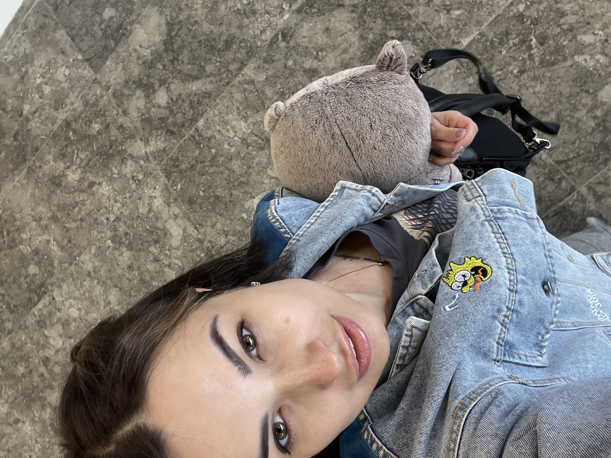 A woman with dark hair and brown eyes looks into the camera, holding a stuffed animal. She is wearing a denim jacket over a dark t-shirt and a necklace. The background appears to be a gray tiled floor.