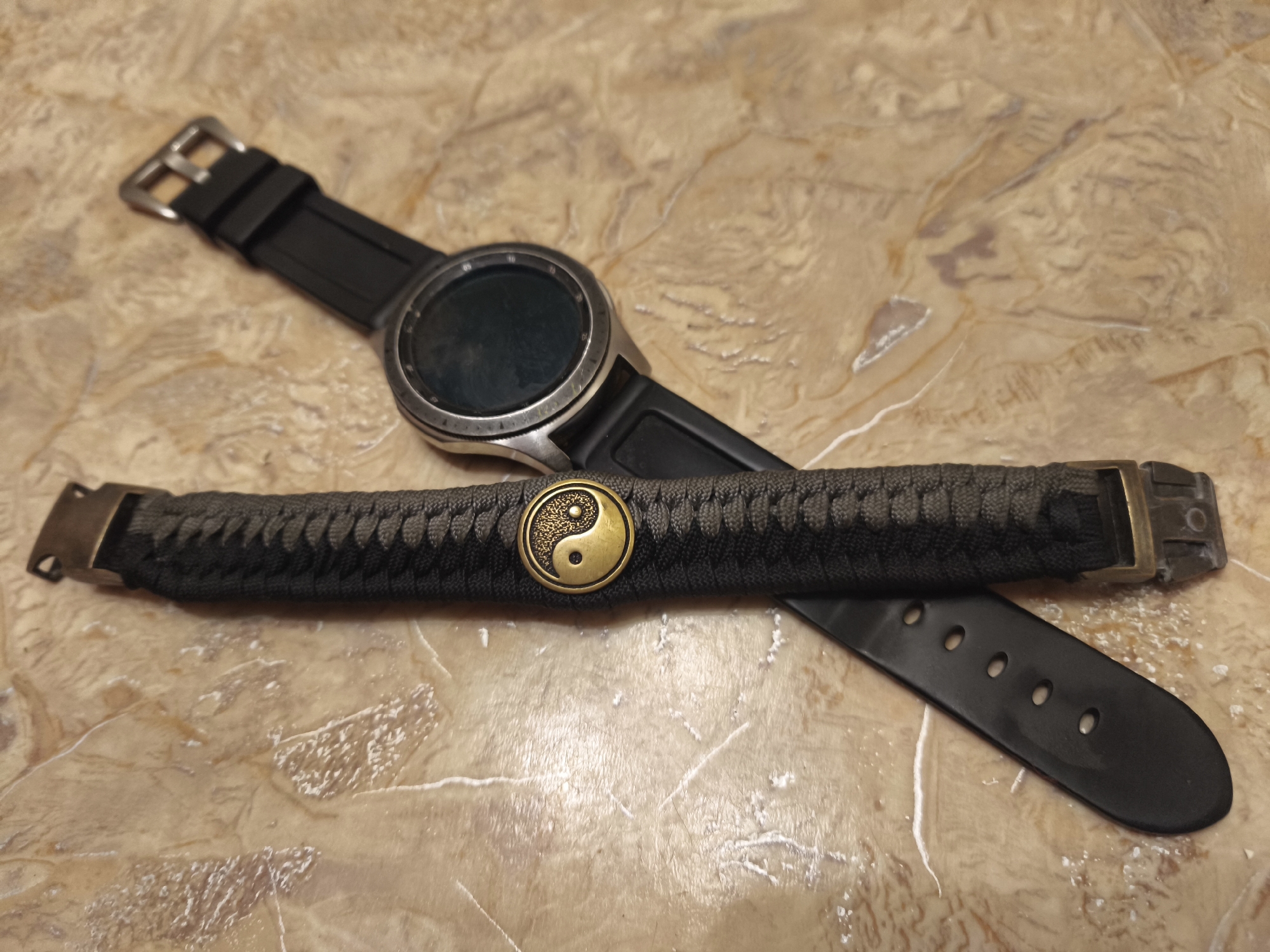 A black smartwatch with a silver bezel is lying on a beige, marble-patterned surface. A paracord bracelet with a yin-yang symbol is resting on top of the smartwatch's band. The bracelet is dark gray and has brass-colored clasps. The smartwatch band is black and has a metal buckle. The surface has a textured, veined pattern. The scene is a top-down view of the objects and surface. There are no people or animals present. The image does not provide any information about the location in the city of Kyiv, Ukraine.