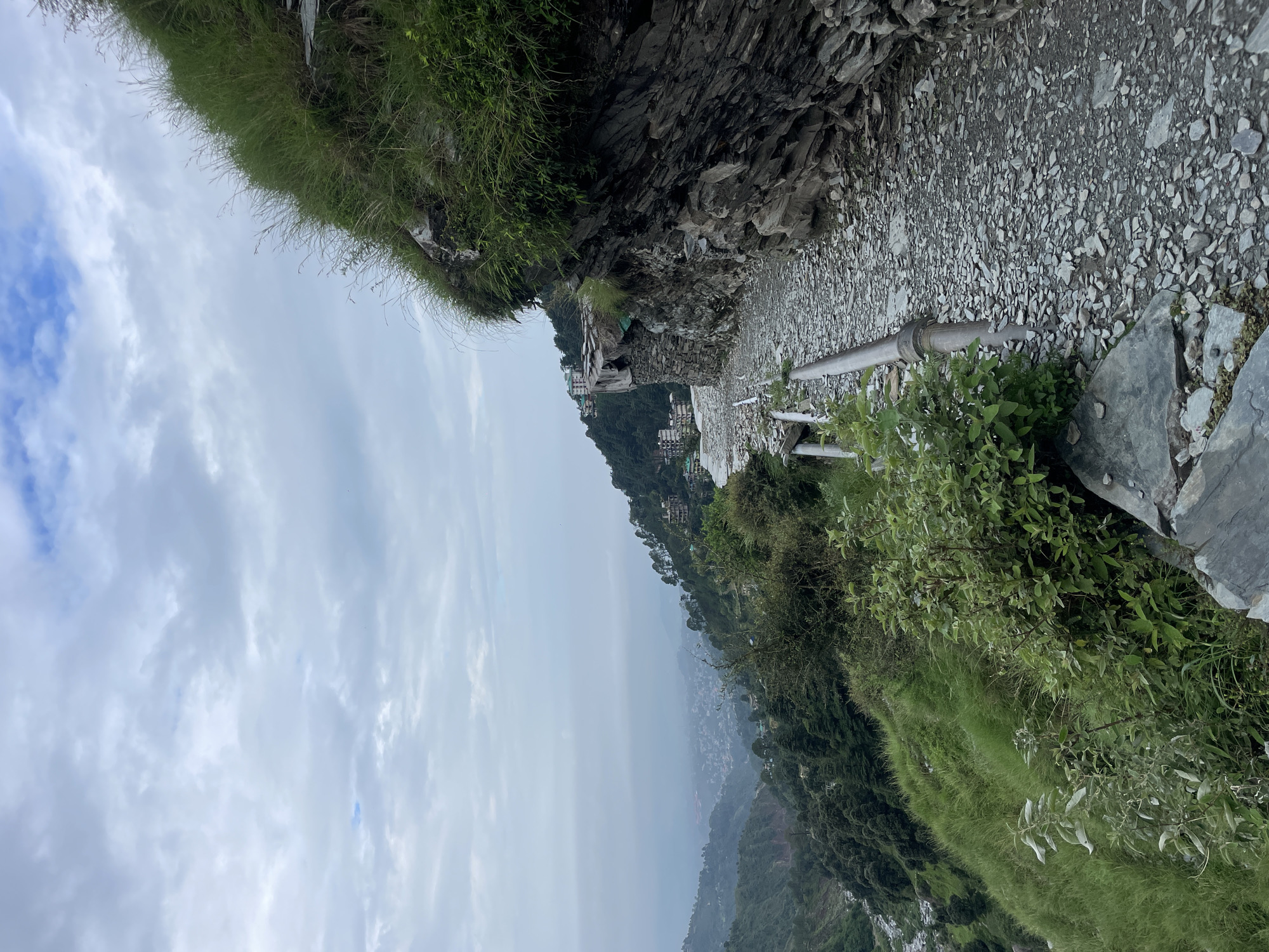 The image depicts a rugged, narrow dirt path winding along a steep mountainside in Bhagsu, India. The path is covered in loose gravel and stones, with a rough-hewn stone wall on the right. A dilapidated metal railing runs along the edge of the path. Lush green grass and bushes grow densely on the slopes of the mountain. In the distance, a hazy valley opens up, revealing a town or village nestled amongst the hills, with buildings and trees visible through the mist. The sky above is overcast with soft, grey clouds. The scene suggests a remote, natural environment with signs of human habitation in the distance. There are no people visible in the image.