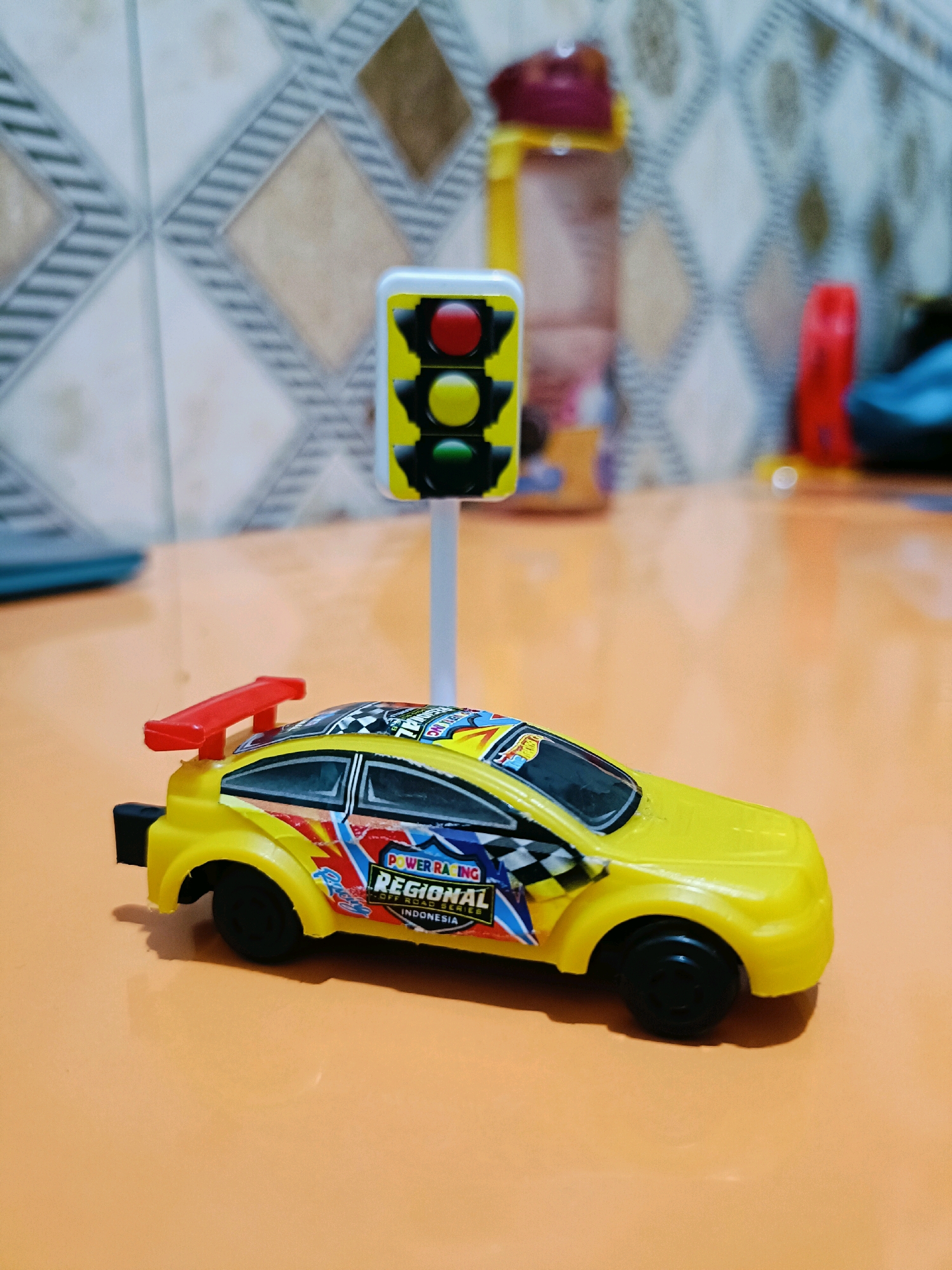 A yellow toy race car with a red spoiler and decals is positioned on an orange surface, with a toy traffic light standing behind it. The background features a tiled wall with a diamond pattern and blurred objects, including a water bottle and a red object. The scene appears to be indoors, potentially a child's play area, and there are no people visible. The location in Candi Wates, Indonesia, is not discernible from the image itself.