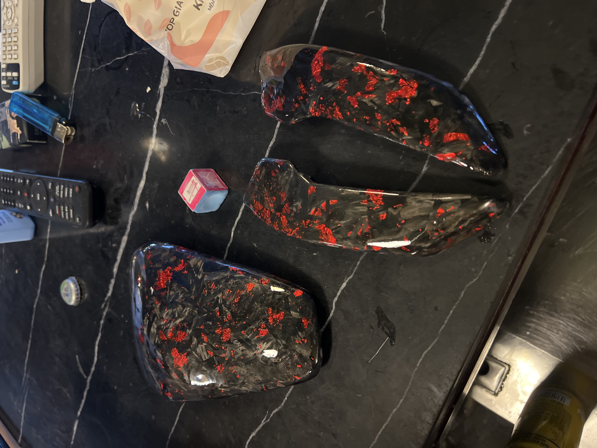 On a black marble countertop in what appears to be a kitchen, lies a collection of seemingly unrelated items. Three pieces of automotive trim with a red and black carbon fiber design are arranged. To the left of the trim pieces, there is a remote control, a blue lighter, a small box with pink and red markings, and a bottle cap. Part of a white package labeled with Vietnamese script, "TOP GIA K..." is visible in the upper left corner. To the right of the trim pieces, a portion of a bottle is visible near the edge of a stainless steel sink. The location in the city of Đà Nẵng, Vietnam, is only based on the Vietnamese writing and there is no additional confirmatory detail.