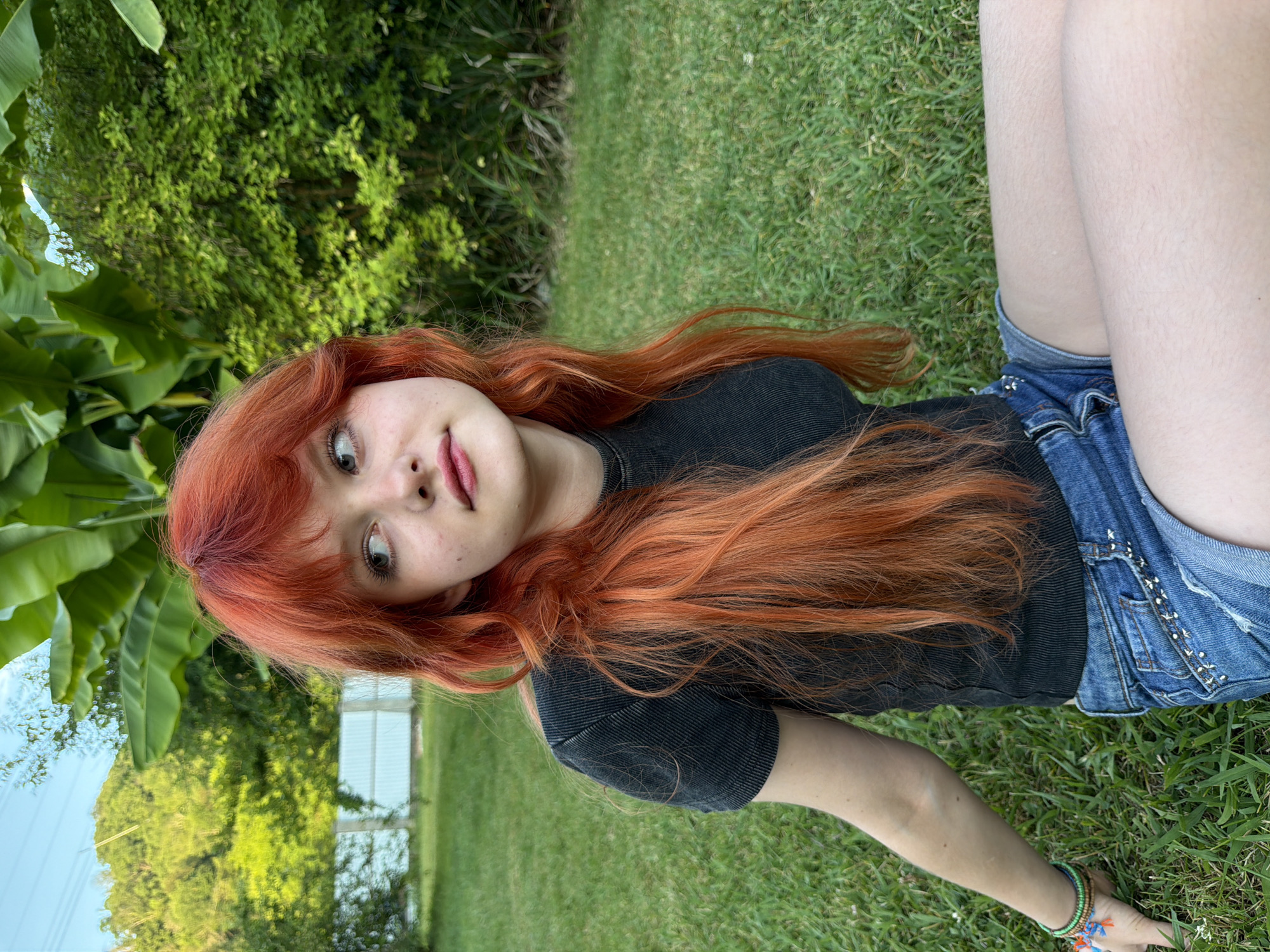 A young woman with long, bright red hair lies on a grassy lawn. She is wearing a dark t-shirt and denim shorts. Her head is tilted, and she is looking towards the camera with a slight smile. Her hair is spread out around her.
The background features lush green foliage and trees, suggesting an outdoor setting. A white building with visible windows can be seen in the distance. The lighting indicates it is daytime, likely late afternoon or early evening, given the soft natural light. The overall mood appears relaxed and casual. The image is oriented vertically.