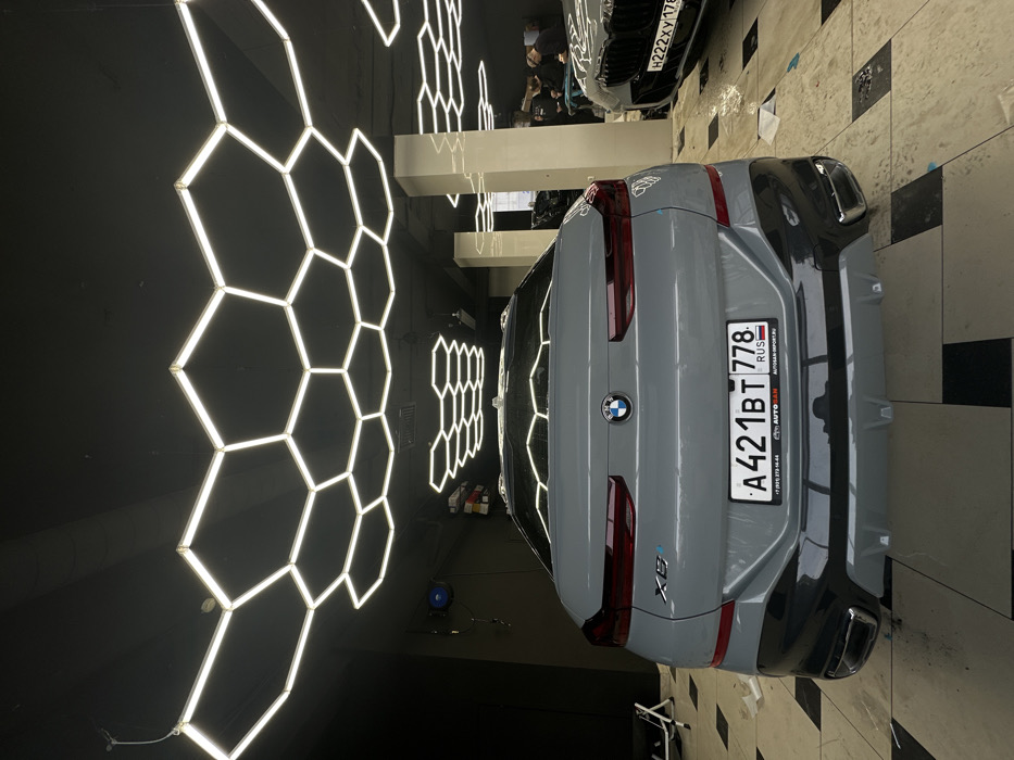 The image depicts an indoor automotive facility in Saint Petersburg, Russia, identifiable by the "778" and "178" region codes on the visible Russian license plates.
The scene features a light grey BMW X6 SUV coupe, viewed from the rear, positioned centrally. The vehicle displays a Russian license plate "A 421 BT 778 RUS" and "X6" and "e" badges on its tailgate. The rear bumper is a contrasting dark grey/black, incorporating chrome exhaust tips. Reflective material, indicative of a protective film or wrap application, is visible on portions of the vehicle's lower body and rear.
To the right, the front end of a second dark-colored BMW, likely an SUV with a prominent black grille, is partially visible. This vehicle also bears a Russian license plate, "H 222 XY 178 RUS". In the background, behind this second vehicle, several indistinct human figures are present, suggesting personnel or customers within the facility.
The environment is characterized by a black ceiling adorned with numerous interconnected hexagonal LED light fixtures, providing bright illumination. The floor consists of large, reflective light and dark grey tiles arranged in a geometric pattern. Dark walls enclose the space, with a blue hose reel mounted on the left wall and white support pillars visible on the right. Additional equipment, possibly a white stool or cart, is located on the far left. The overall impression is that of a modern automotive detailing or service shop where vehicles are undergoing work.