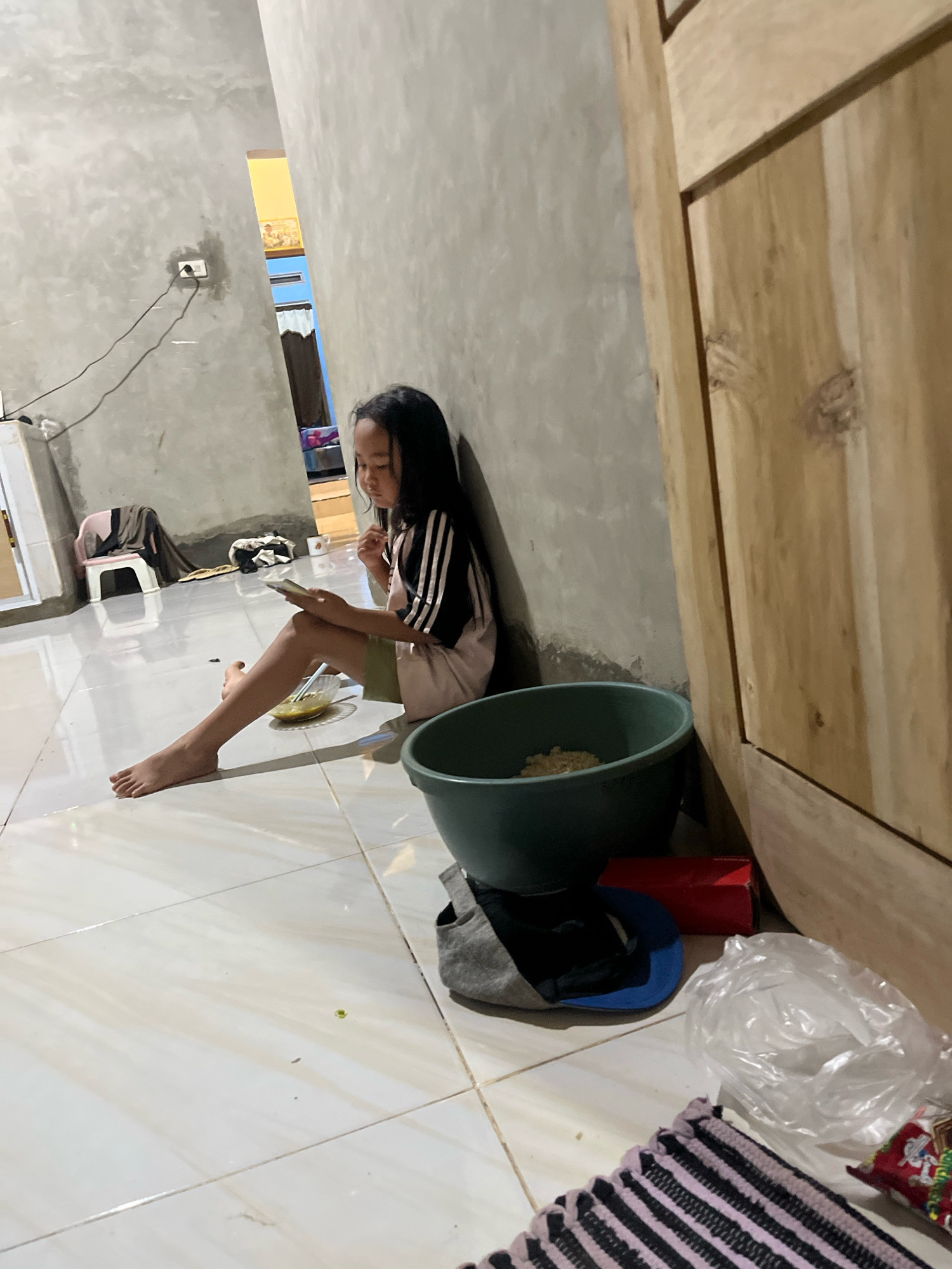 A young girl sits on a tiled floor, leaning against a gray, textured wall. She has long, dark hair and is wearing a pink t-shirt with black stripes on the sleeves and shoulders. Her legs are extended in front of her, and she is holding a phone in her left hand while looking down at it. To her right, there is a small bowl of food with a spoon. A large, dark green basin containing some light-colored food is visible next to her.
In the background, a wooden door or cabinet is on the right side of the image. The wall has an electrical outlet with a black cord plugged in. Further back, there's a glimpse of another room with yellow and blue walls, and curtains. A small pink stool with a draped cloth is in the mid-ground.
On the floor near the basin, there are various objects including a grey cap with a blue brim, a red box, a crumpled plastic bag, and a striped mat. A small green speck is on the floor in the foreground. The lighting suggests it might be evening or indoors. The scene appears to be a domestic setting in Kebumen, Indonesia. The girl seems focused on her phone, possibly eating or taking a break.