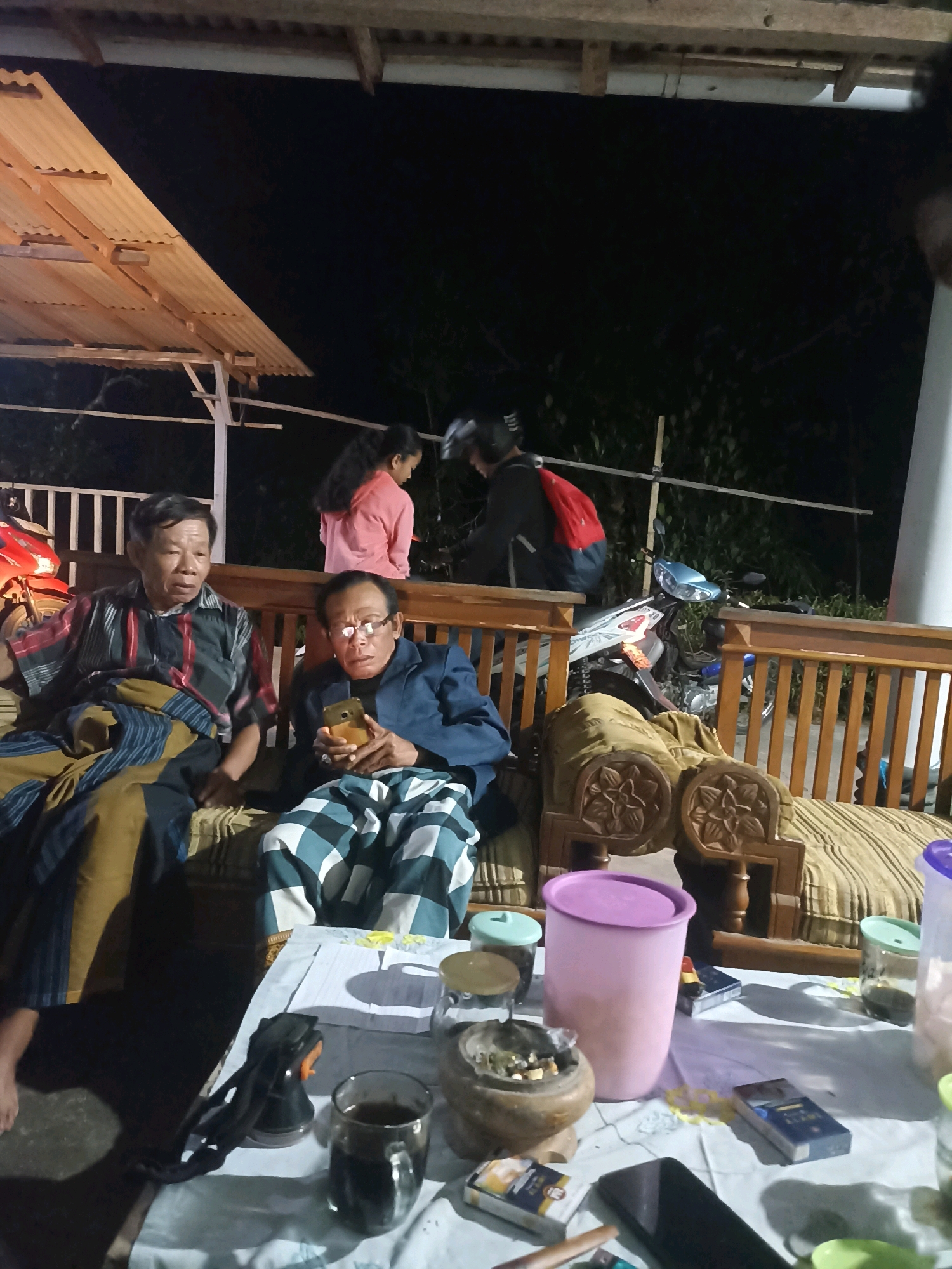 The image depicts a dimly lit outdoor scene, likely in Wonogondo, Indonesia, at night. Two men are seated on a wooden bench with a striped cushion. The man on the left wears a patterned shirt and a sarong, while the man on the right wears a dark blue jacket and glasses, holding a smartphone. In the background, a young girl in a pink shirt stands, and a person wearing a helmet and a red backpack is seated. Several motorcycles are visible behind them, parked near a wooden fence and foliage. A table in the foreground is covered with a white cloth and holds various items, including a glass of dark liquid, an ashtray with cigarette butts, packets of cigarettes, a pink plastic container, and glass jars. The overall atmosphere suggests a casual gathering or rest stop.