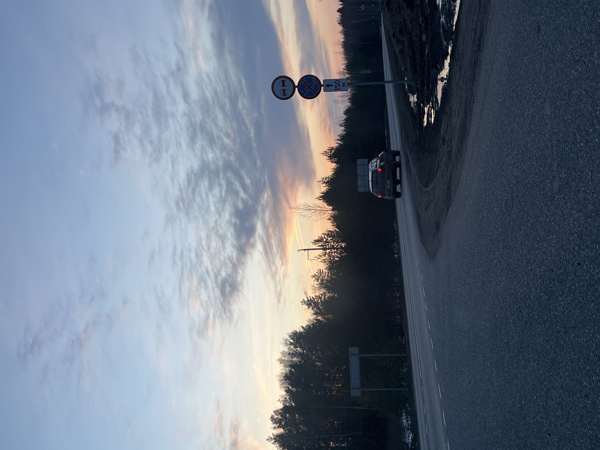 The image depicts a road in Chaikovsky, Russia. A silver SUV is driving on the right lane of the road, away from the camera. On the left side of the road is a dense forest. Road signs are visible on the left side of the road, including "No Stopping" and "No Parking" signs, with a distance marker indicating 150 meters. The sky is visible above the trees, displaying a sunset with a mix of blue, orange, and pink hues. The road is asphalt.
