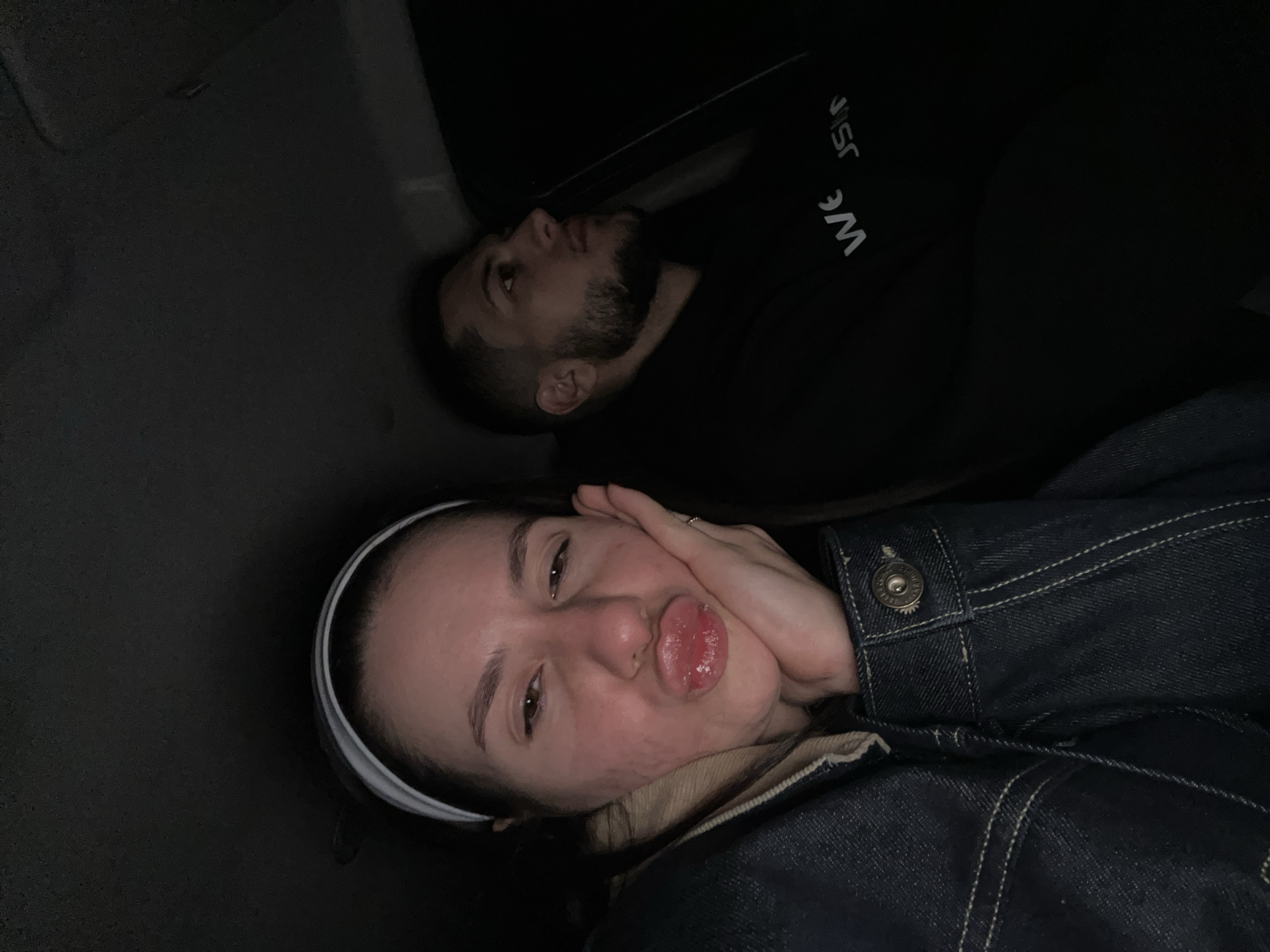 The image shows two people lying down. A woman is in the foreground, wearing a denim jacket and a white headband, making a pouting face. A man with a beard is lying behind her, wearing a black top with white lettering. The setting appears to be inside a vehicle, possibly a car. The background is dark and indistinct. The location in the city of Yastrebovo, Bulgaria is not discernable.